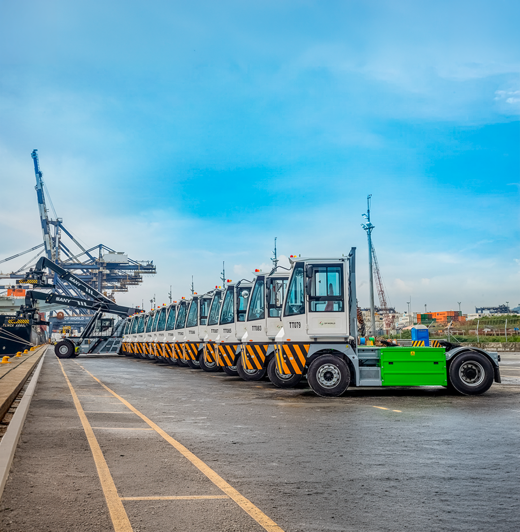 Row of electric terminal tractors parked at DP World in Santos port terminal with cargo handling equipment and cranes in the background.