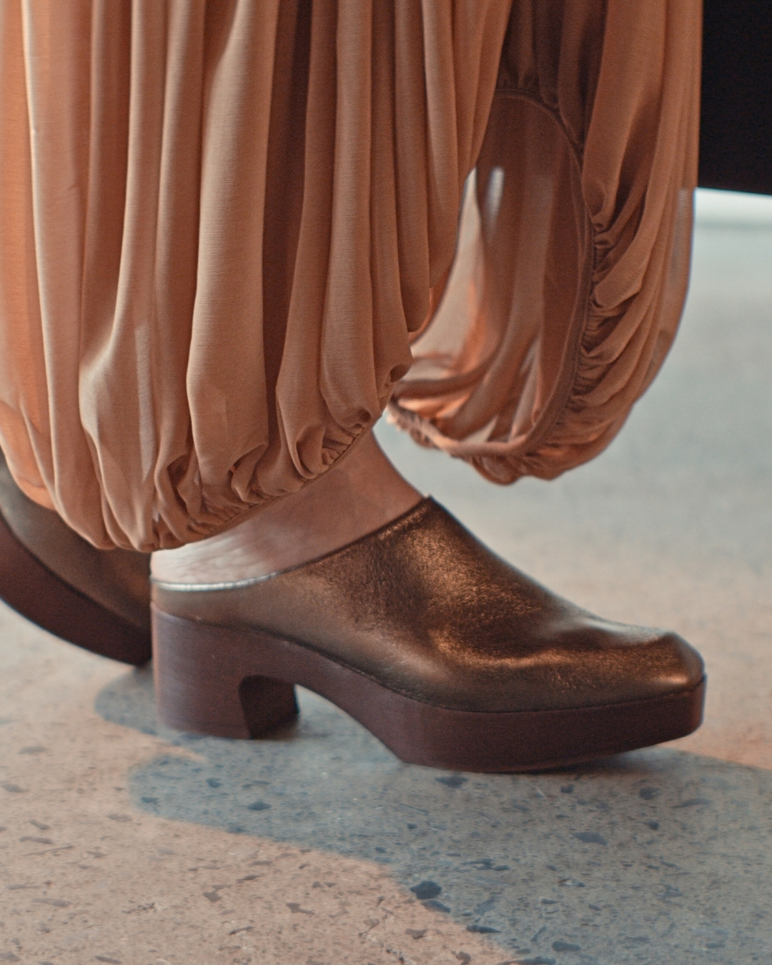 Close-up of feet wearing bronze metallic clogs with light wood platform soles and block heels beneath gathered trousers.