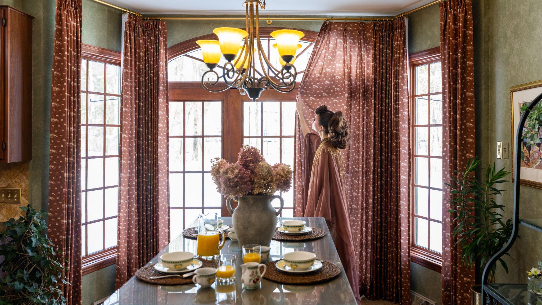 Ms. Maverick Muse adjusting a window curtain in a dining area, featuring TWOPAGES Print Linen Cotton Drapery in Dusty Clay MM-F03 pattern.