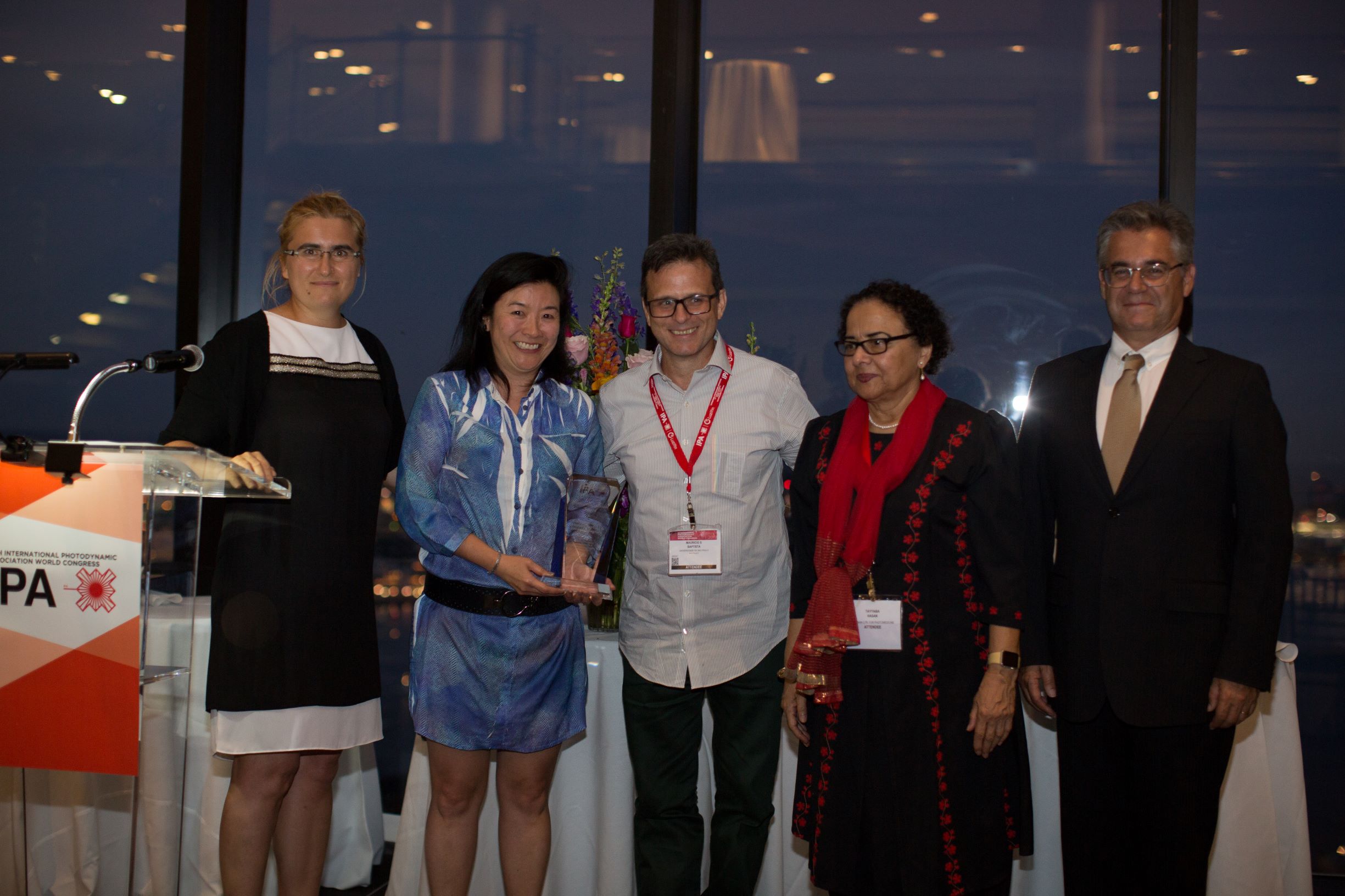 Professors Cristina Kurachi (center-left) and Mauricio Baptista (center) receiving the 2019 Humanitarianism Award from Patycja Nowak-Sliwinska (left), IPA Past-President, Dr. Tayyaba Hassan (center-right), and IPA President, Dr. Luis Arnaut (right). Professor Vanderlei Bognato not pictured.