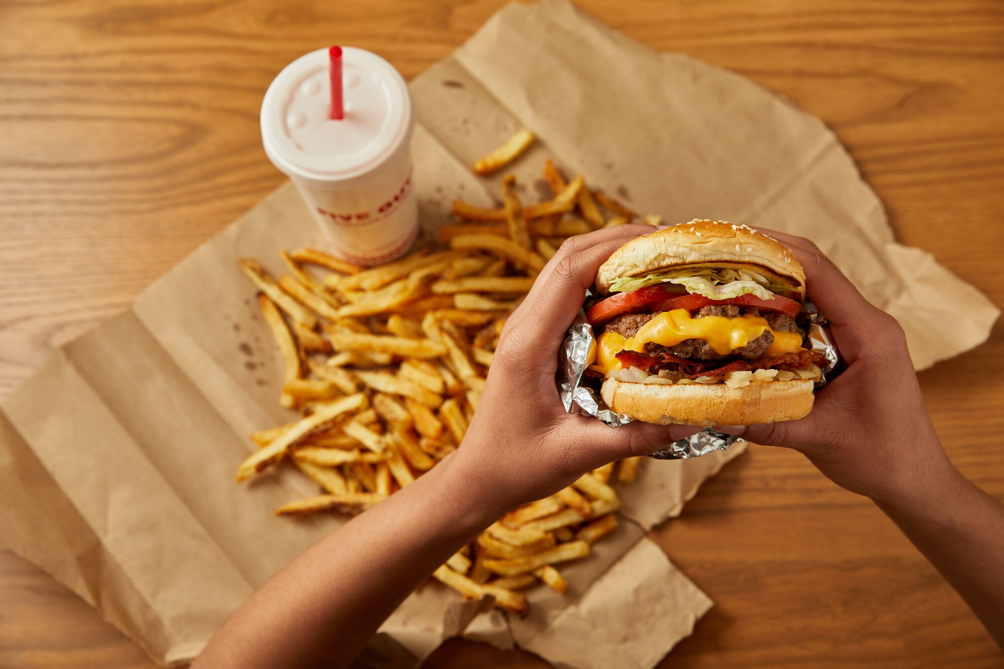 Hands holding a Five Guys cheeseburger over fries, with a drink on a wooden table.