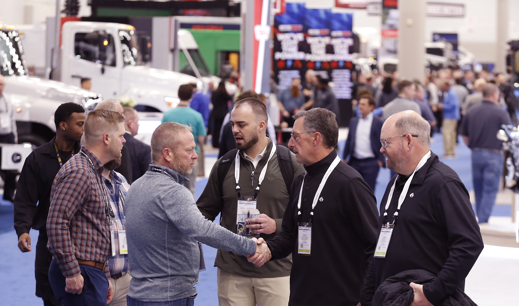 Two men in a group of six shake hands on a busy commercial vehicle trade show floor with trucks and crowd in the background. 
