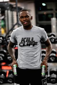 UFC Champion Israel Adesanya wears a Kill Cliff shirt and holds Kill Cliff drink cans standing in a gym
