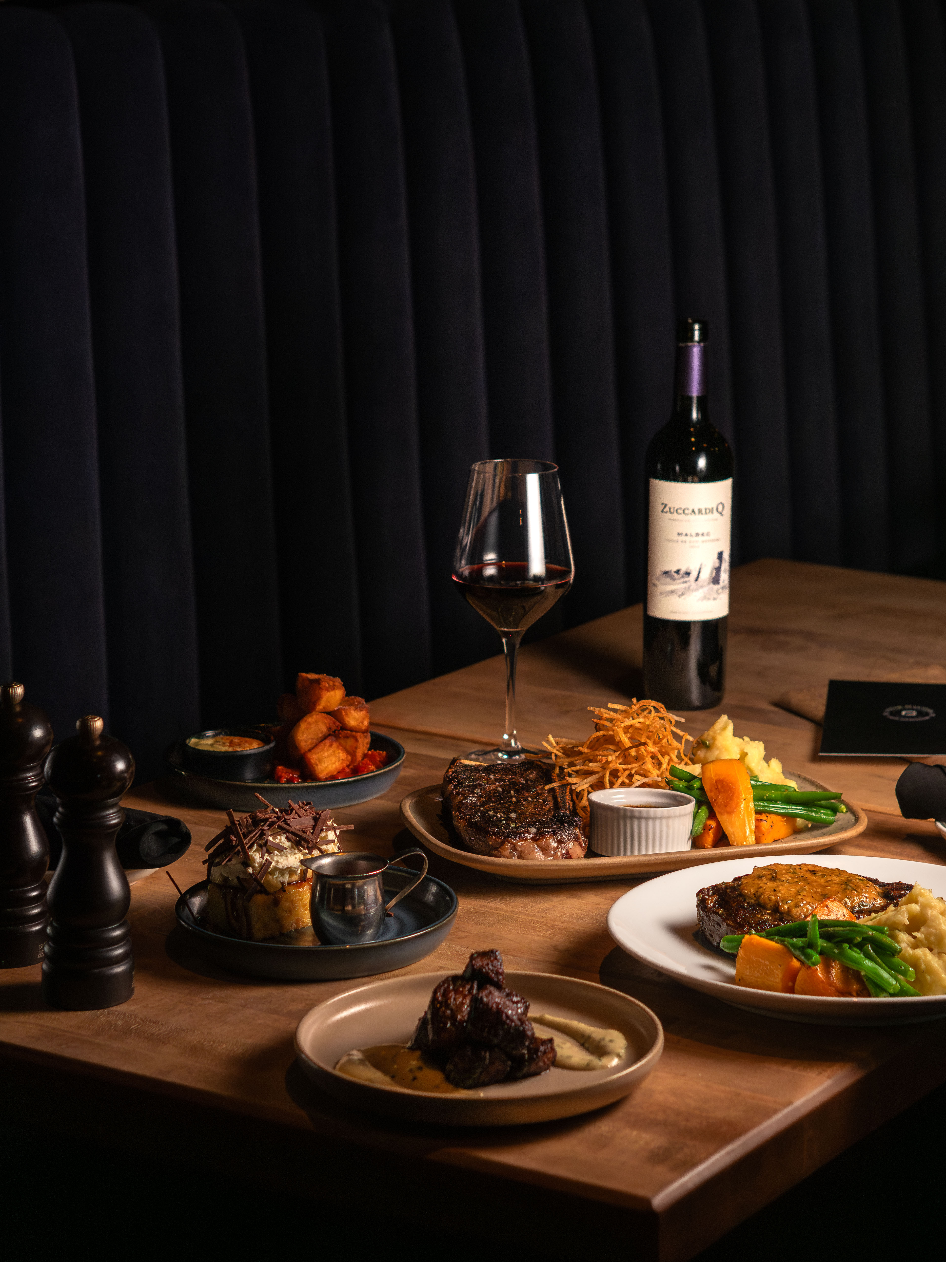 A table with several different dishes including a bottle of red wine, a banoffee bread pudding desert and steak main dishes