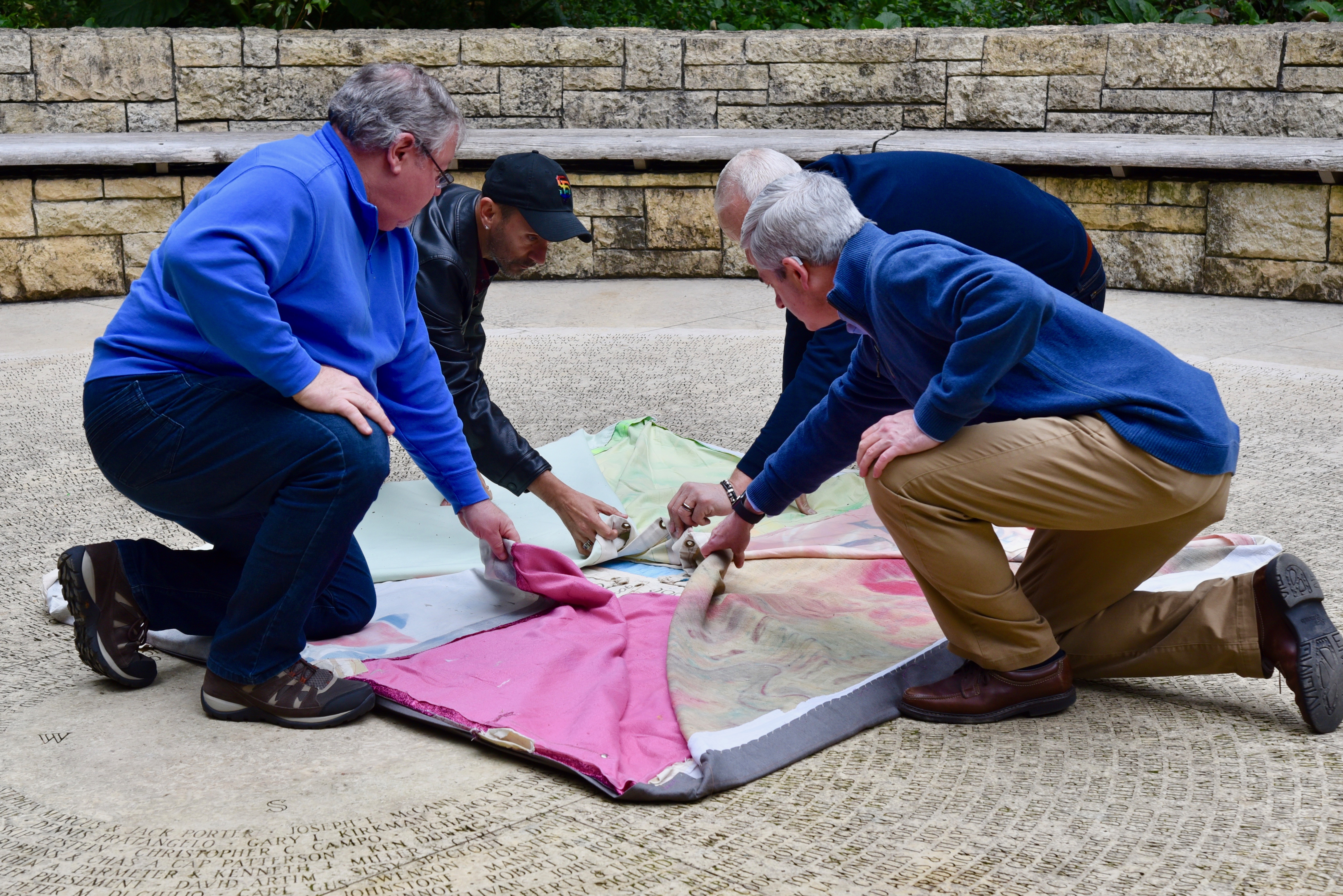 Photo 1: The first AIDS Quilt panels to arrive back in San Francisco is ceremoniously unfolded in the Circle of Friends at the National AIDS Memorial. It is one of 25 blocks of panels that will be on display during 2019 World AIDS Day ceremonies at the National AIDS Memorial. All 50,000 panels of the AIDS Quilt will permanently move from Atlanta to San Francisco in 2020 under the care and stewardship of the National AIDS Memorial.   Photo Credit: Mike Shriver.  (L to R) Mike Smith, one of the founders of the AIDS Memorial Quilt; Kelly Hart, National AIDS Memorial AIDS Quilt Coordinator; John Cunningham, Executive Director, National AIDS Memorial; Brett Pletcher, Executive Vice President, Corporate Affairs and General Counsel, Gilead Sciences.  