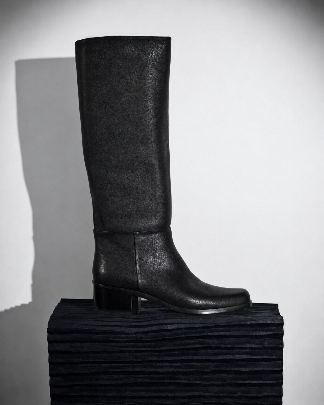 Black knee-high leather boot with a square toe and low stacked heel displayed in profile on a black sculptural pedestal.