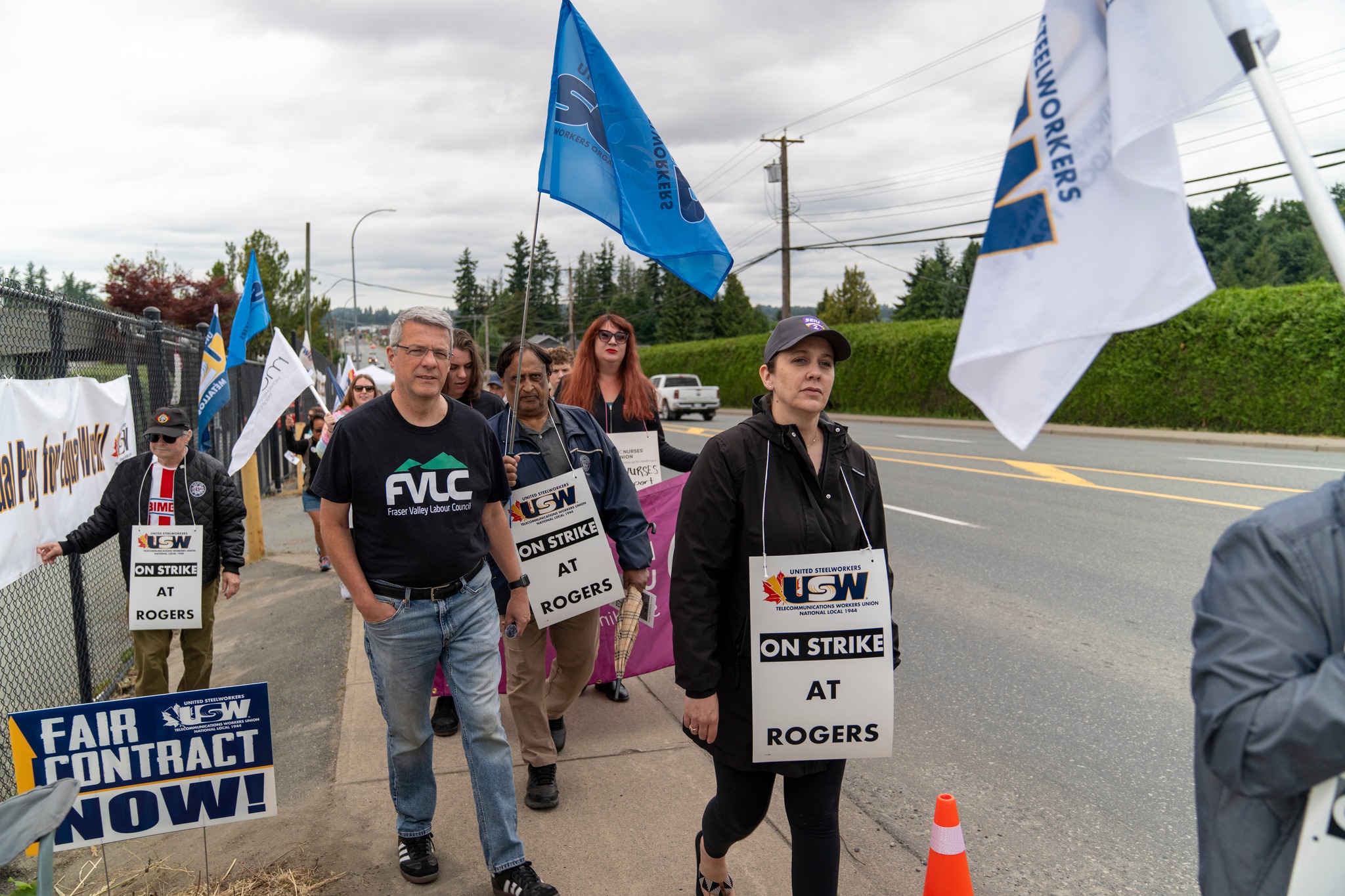 A group of USW Local 1944 union members march on a sidewalk during a strike, holding signs that read "USW ON STRIKE AT ROGERS." Some of the strikers a