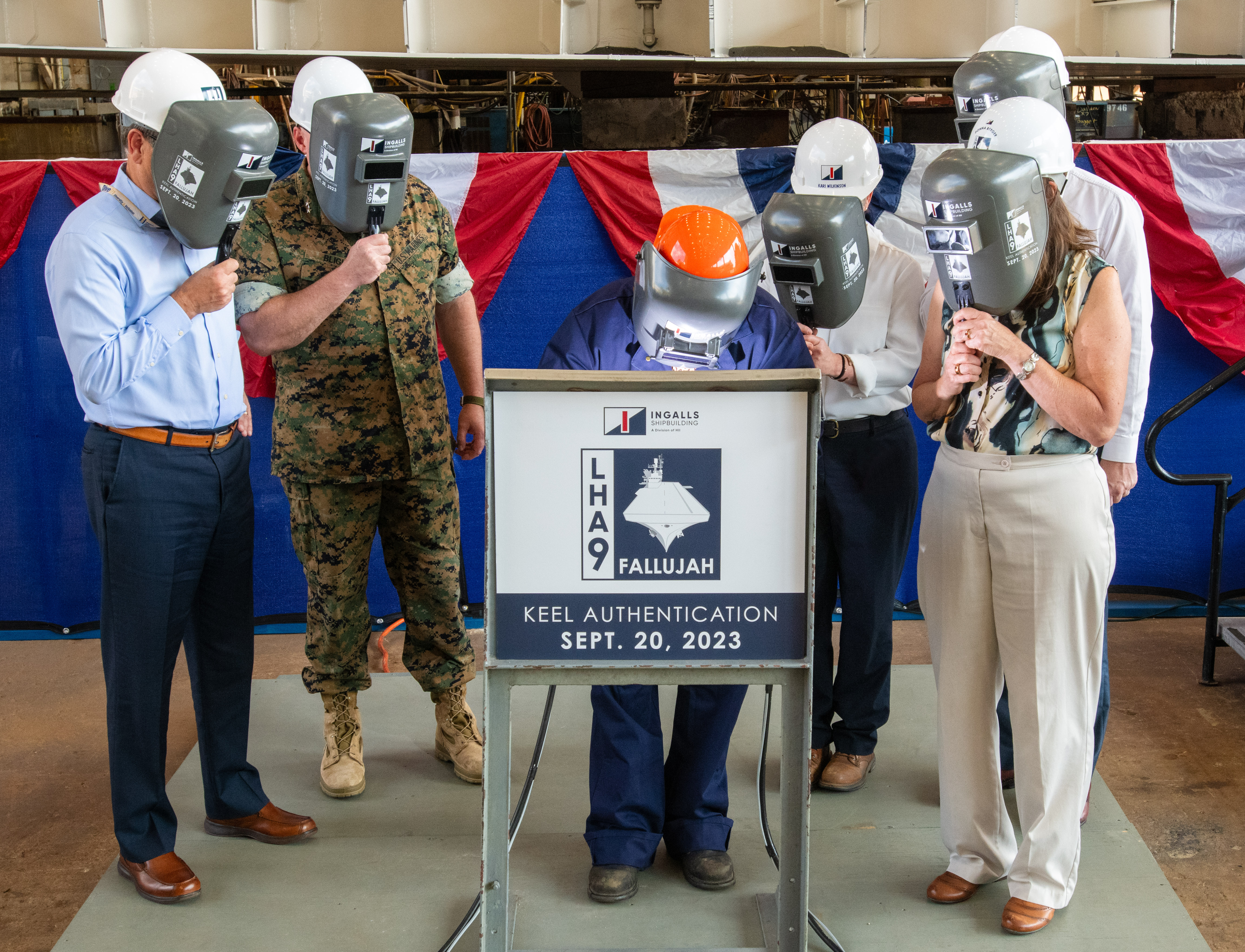 Ingalls welder Seveta Gray welds the initials of Donna Berger onto the keel plaque that will be permanently part of Fallajuh (LHA 9).  Left to right, HII President & Chief Executive Officer Chris Kastner, U.S. Marine Corps Maj. Gen. David Bligh, Ingalls Shipbuilding President Kari Wilkinson, Under Secretary of the Navy Erik Raven, and Ship Sponsor Donna Berger.