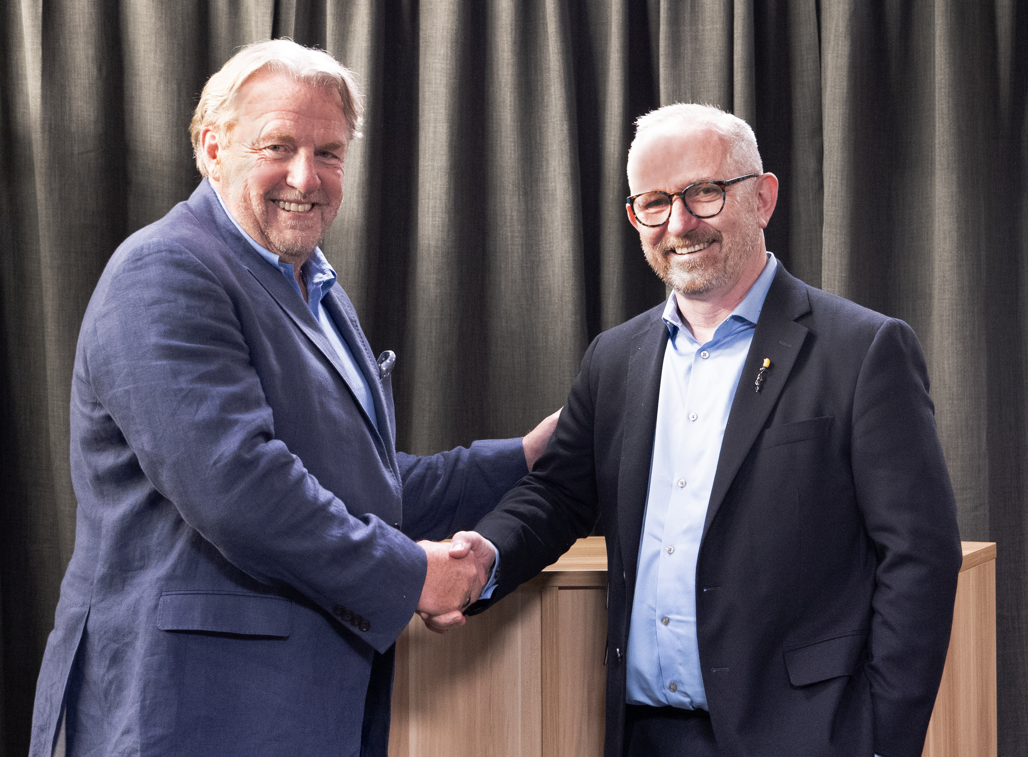 Two men in suits smiling and shaking hands in front of a dark curtain, symbolizing the signing of a partnership agreement