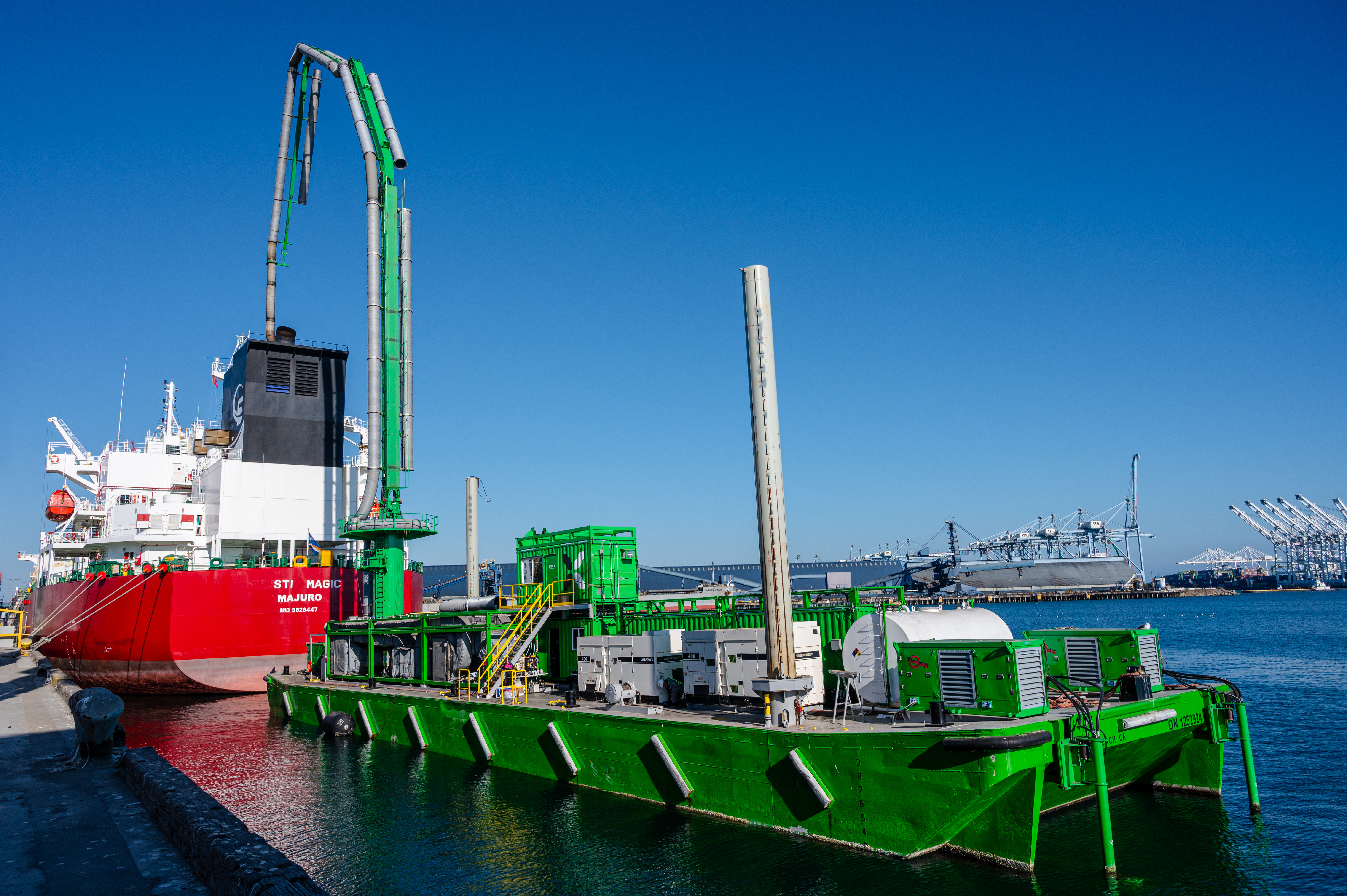 Bright green STAX barge connected to the exhaust pipes of a large red and white tanker vessel in a port on a clear blue-sky day.