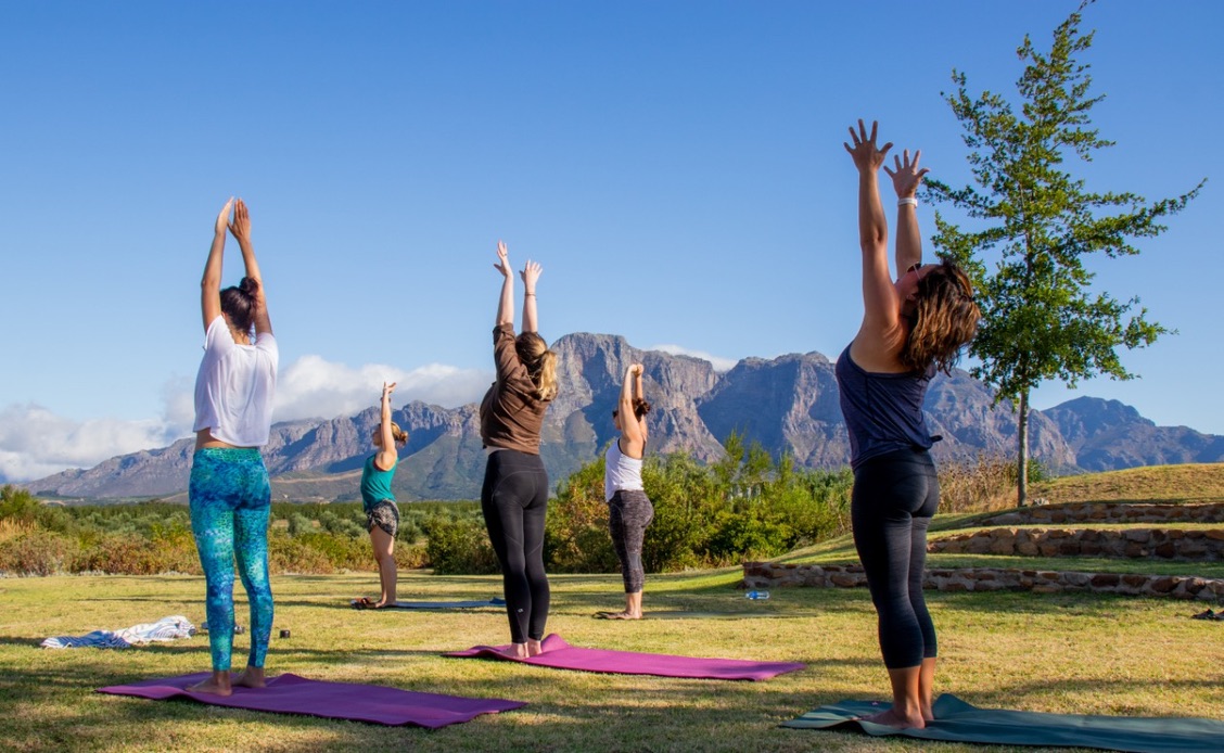 Women practicing yoga on grass with mountains of the Cape Winelands in the background under a clear blue sky.