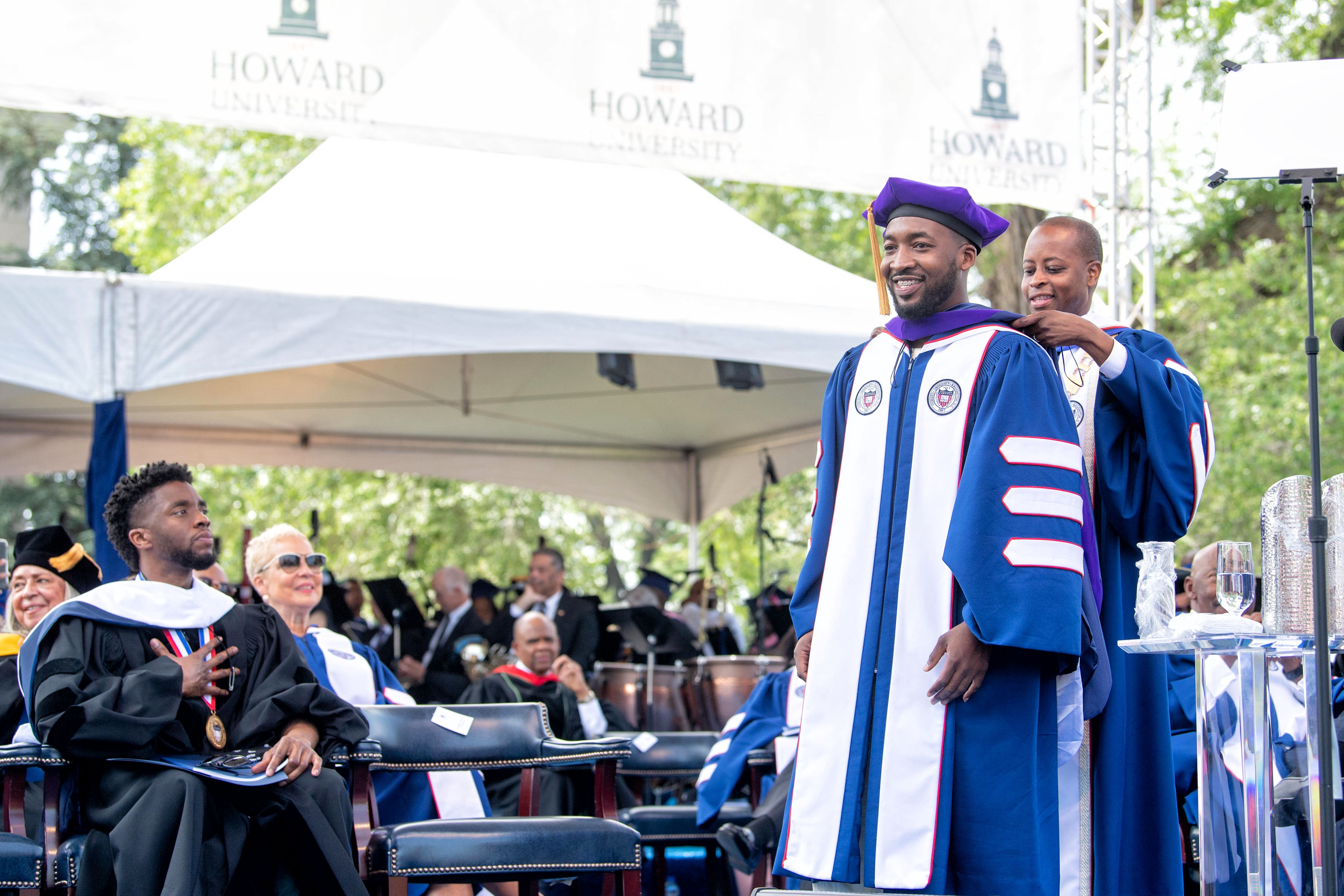 President Wayne A. I. Frederick hoods graduate during the 2018 commencement celebration as speaker Chadwick Boseman looks on.  