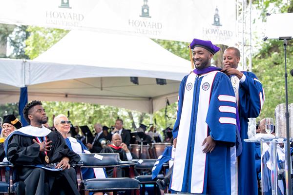 President Wayne A. I. Frederick hoods graduate during the 2018 commencement celebration as speaker Chadwick Boseman looks on.  