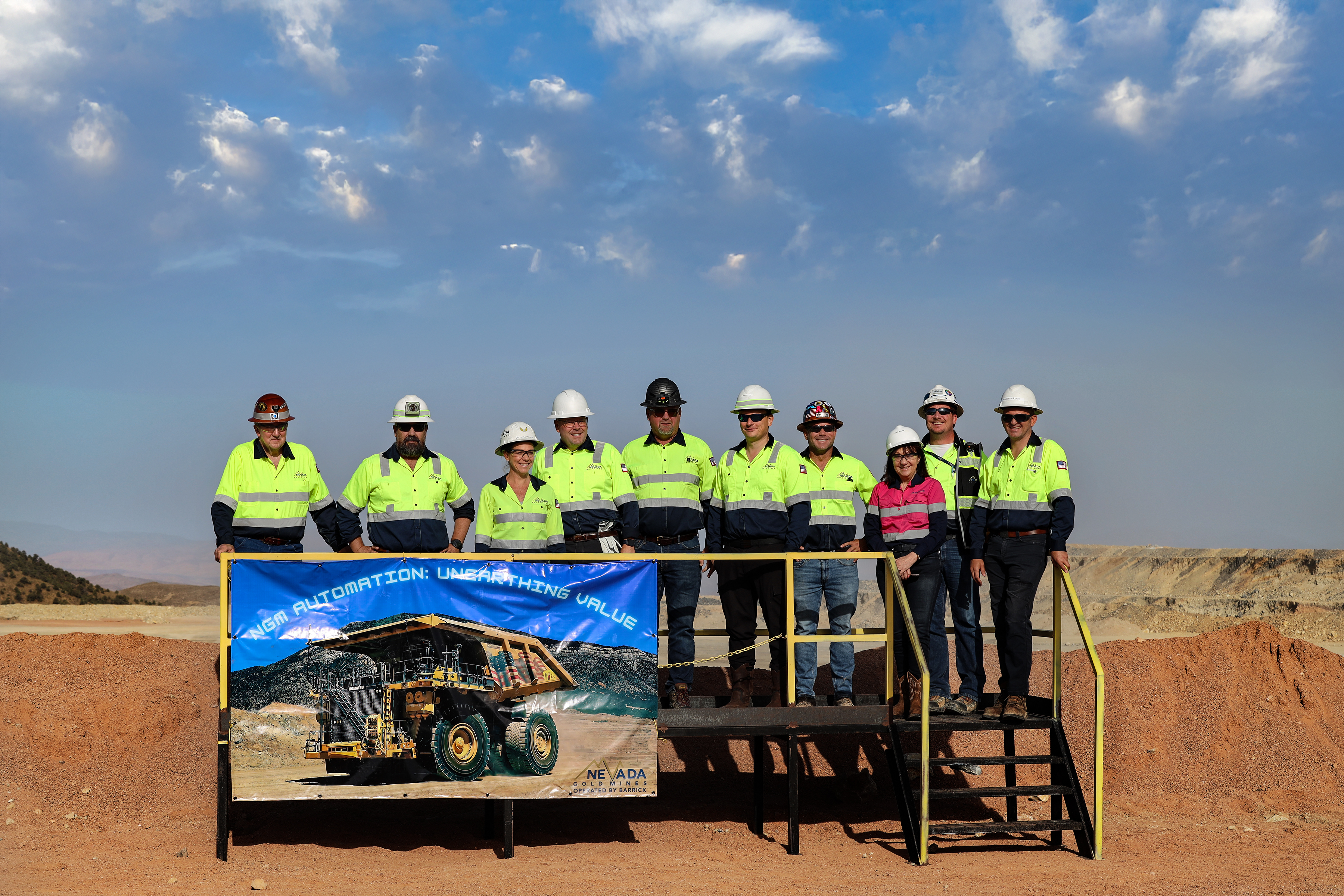 A group of ten mining workers in bright yellow high-visibility safety shirts with reflective stripes and hard hats pose on a yellow metal platform.