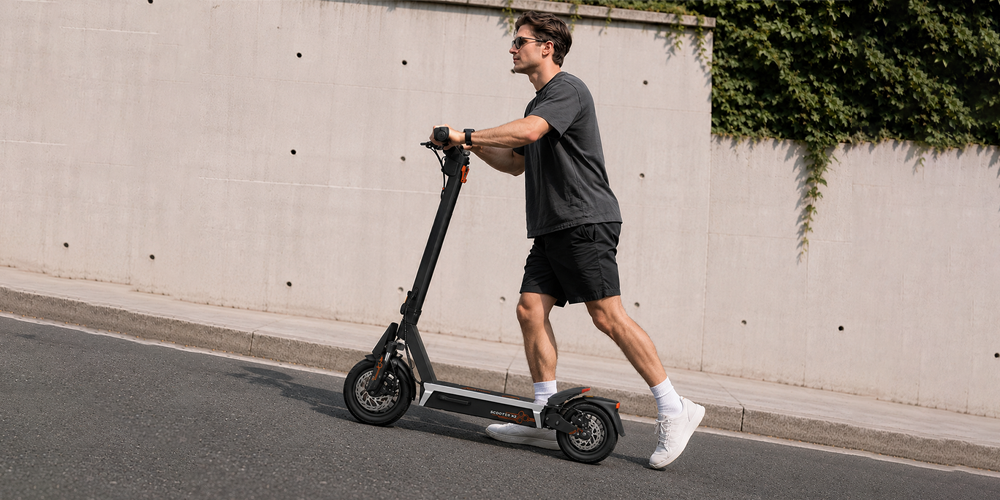 Young man pushes lightweight urban ONECNA T2 electric scooter along city pavement beside concrete wall