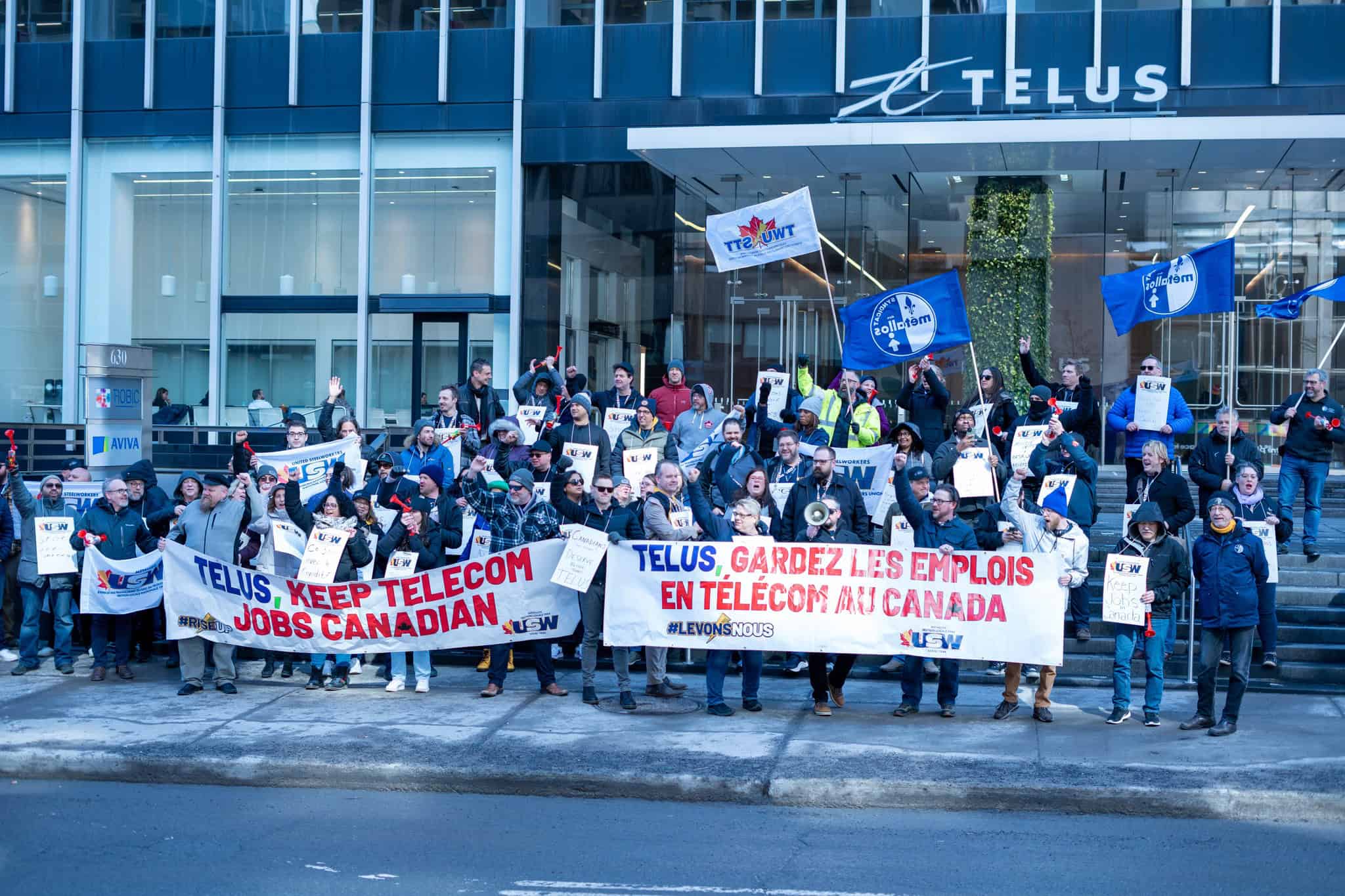 Image: A group of workers hold banners outside a Telus building, saying "Telus: Keep Telecom Jobs Canadian."