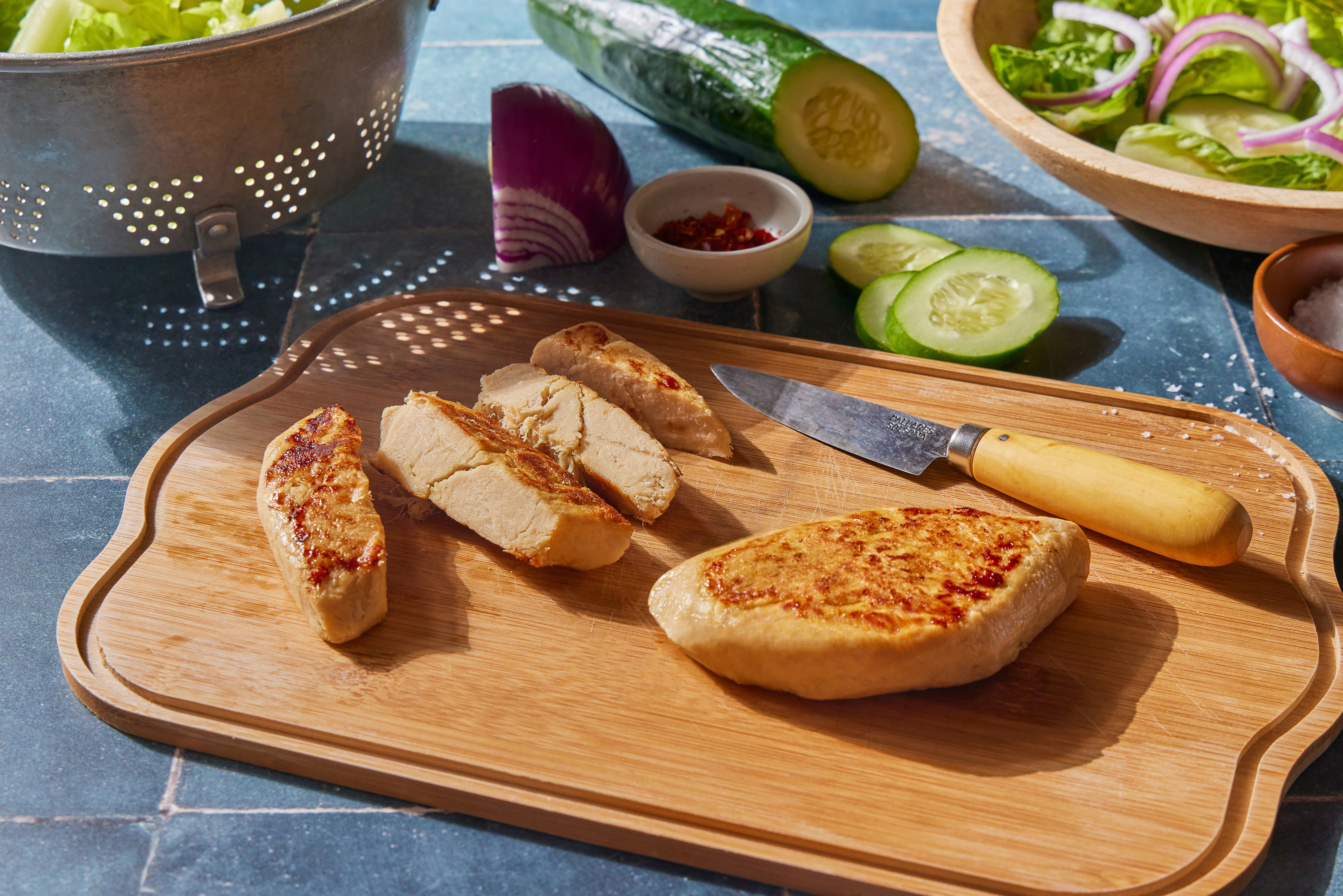 A wooden cutting board with sliced and whole pieces of Tender plant-based chicken breast. A knife with a wooden handle rests beside the chicken.