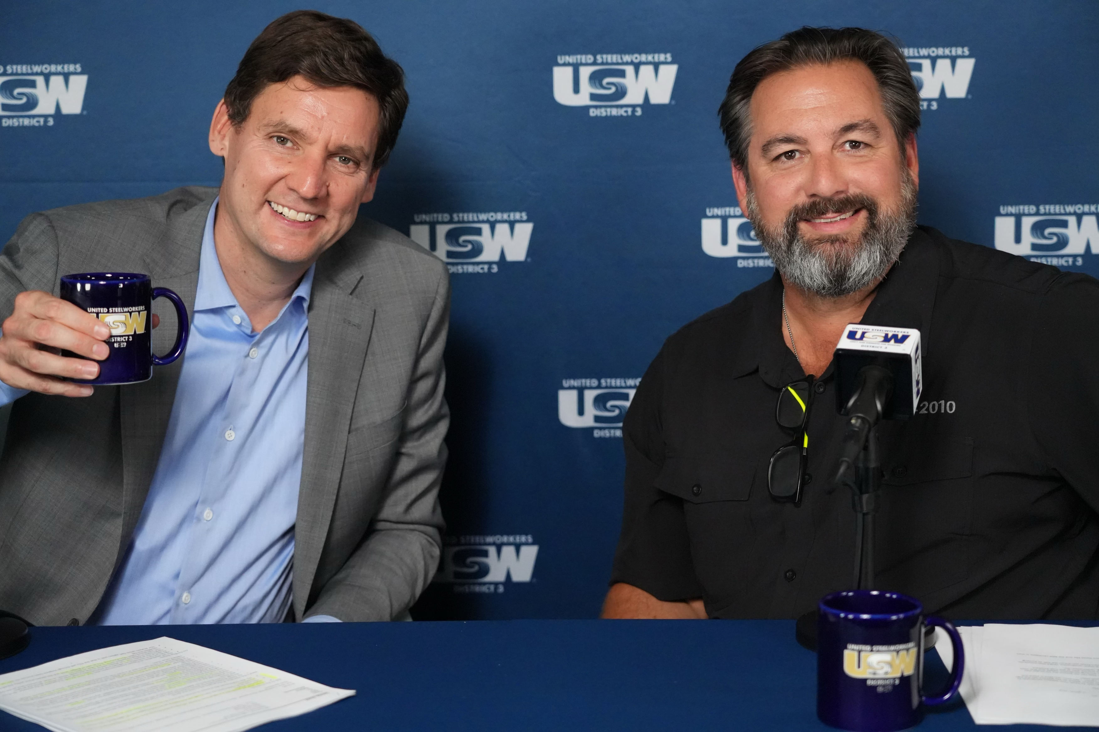 Image: Two people are seated at a table facing the camera, smiling. The one on the left is wearing a suit jacket and dress shirt (no tie) and raising 