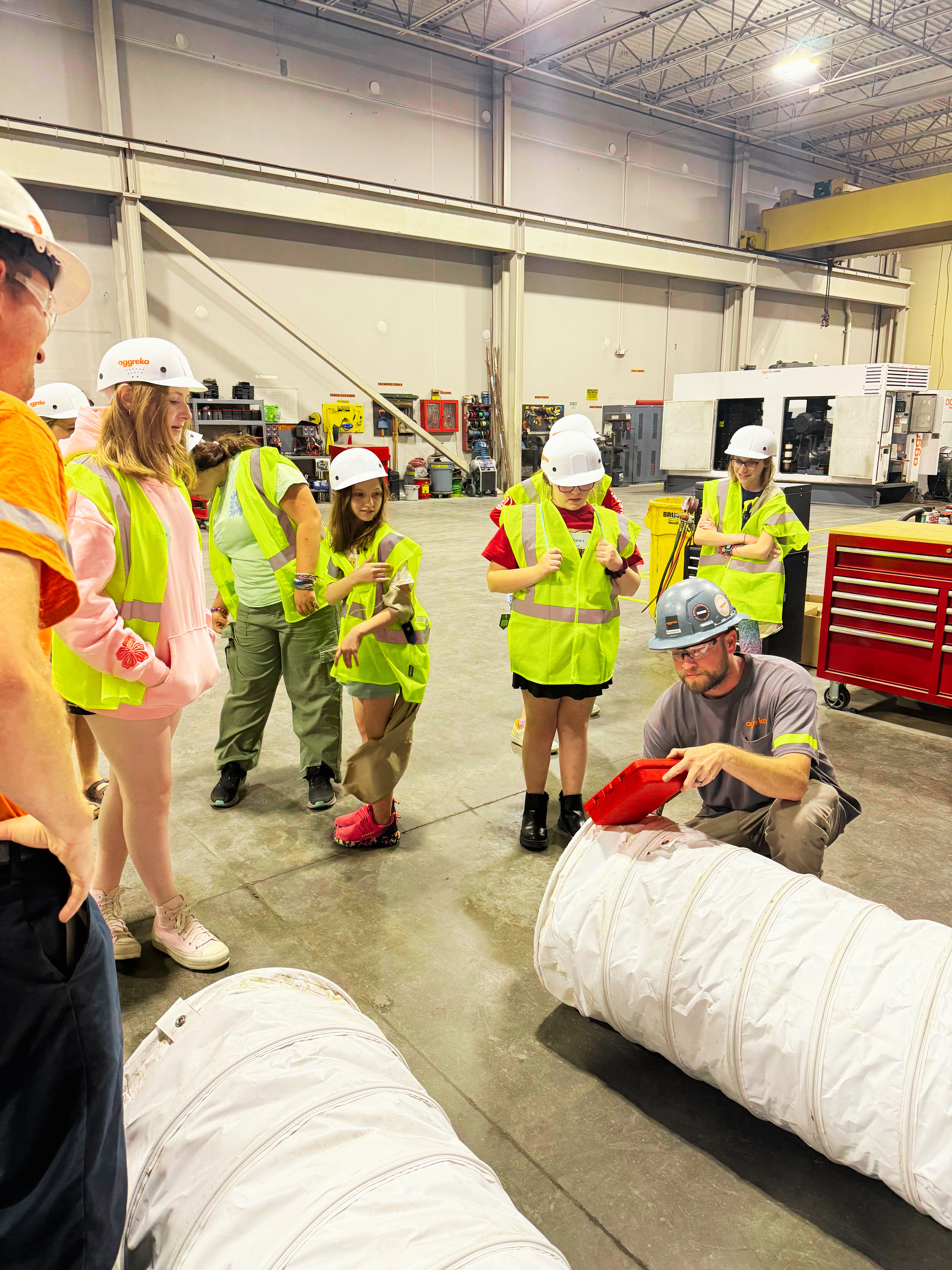In a warehouse, a group of girls in safety vests and hardhats observe an engineer displaying a toolbox