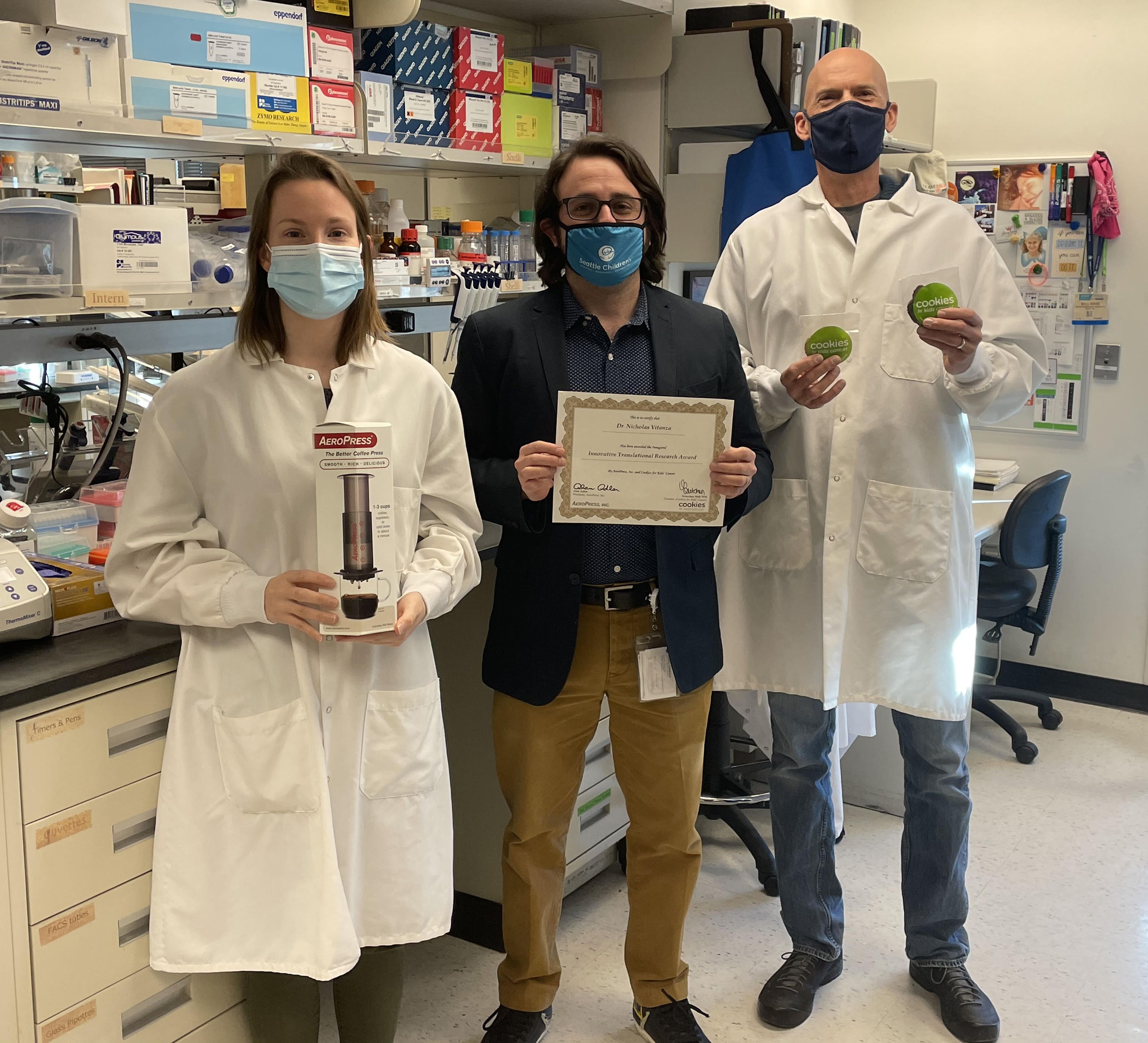 Palo Alto, CA - Dr. Nicholas Vitanza (center) of Seattle Children's Research Institute is the winner of the $100,000 inaugural Innovative Translational Research Award underwritten by AeroPress, Inc. and administered by Cookies for Kids' Cancer. Dr. Vitanza and his fellow lab founders, Senior Research Technician Carrie Myers (left) and Senior Research Scientist Matt Biery (right), are pictured here with an award certificate prize pack including an AeroPress coffee maker and fresh-baked cookies from Cookies for Kids' Cancer. Dr. Vitanza is a pediatric neuro-oncologist at Seattle Children’s researching midline gliomas, a particularly aggressive form of brain tumor. His hope is to develop an immunotherapy with chimeric antigen receptor T cells to treat children who typically have a survival of only 11 months after diagnosis.