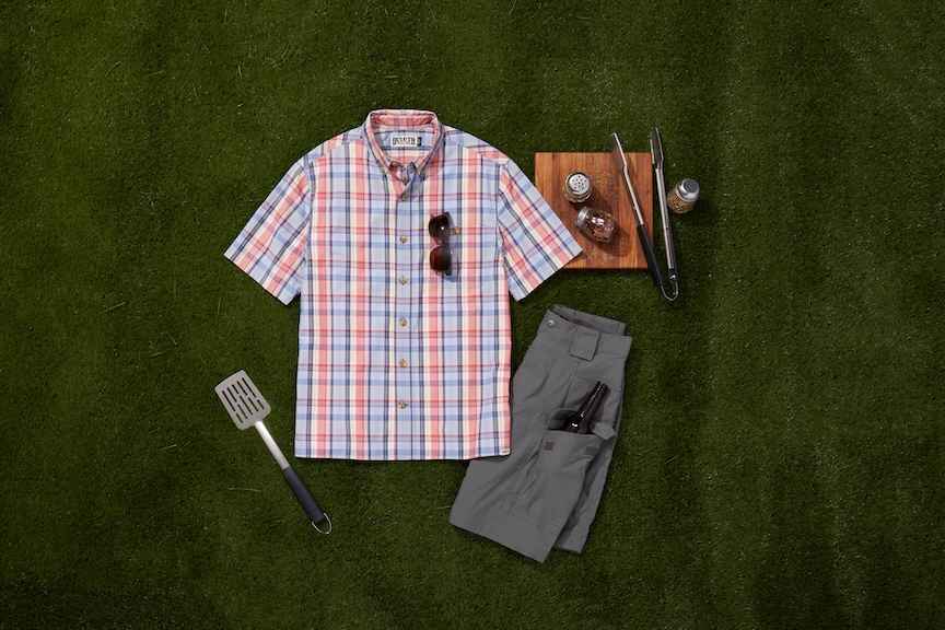 Duluth Trading Co. BBQ collection featuring a plaid short-sleeve shirt, gray grilling shorts with beverage pocket and grilling tools on green grass.