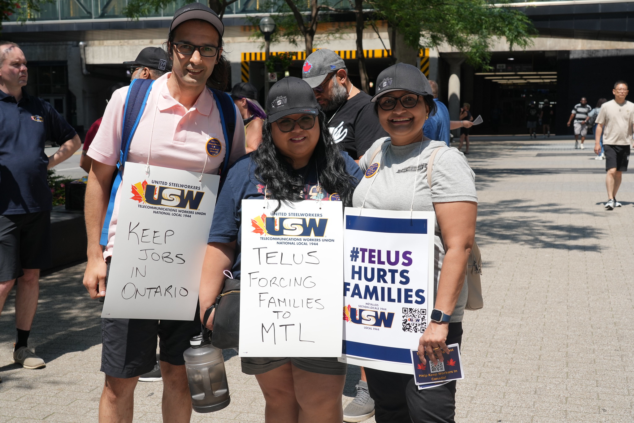 Image: three people stand outdoors on a sunny day wearing placards: "#Telus hurts families" and "Keep jobs in Ontario."