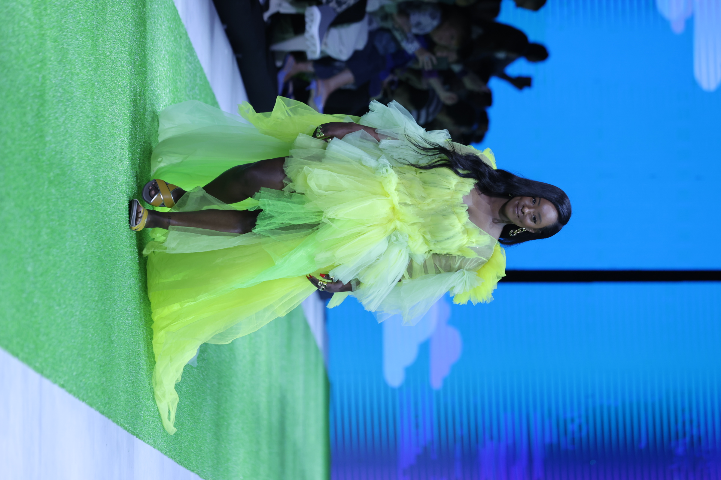 Image shows model on a runway wearing a neon green tulle dress with Naot sandals