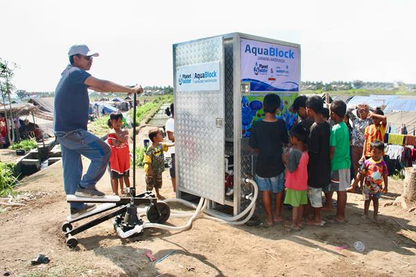 Families are able to drink clean, safe water from a Planet Water AquaBlock located in a relocation camp in Lombok, Indonesia after a series of earthquakes destroyed water and electric systems in August, 2018. The AquaBlock features three Xylem pumping solutions, whether it be petrol, electric or in manual mode using a stepper pump (shown in use). Recently, the emergency response drinking water kiosks have been successfully deployed in Africa (Cyclone Idai) and relief camps India (floods). 
