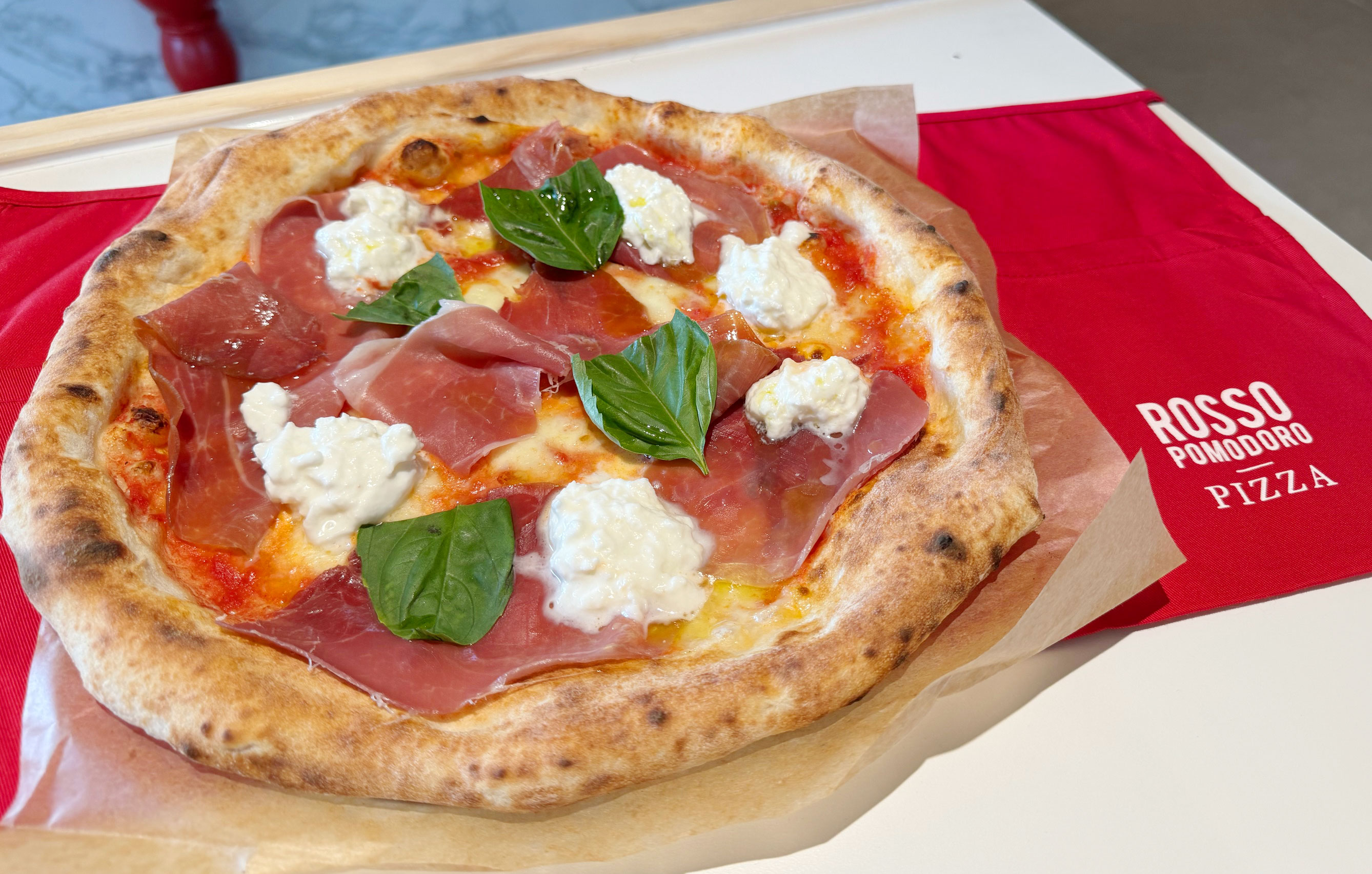 Image shows a Neapolitan style pizza from Rosso Pomodoro with prosciutto and burrata