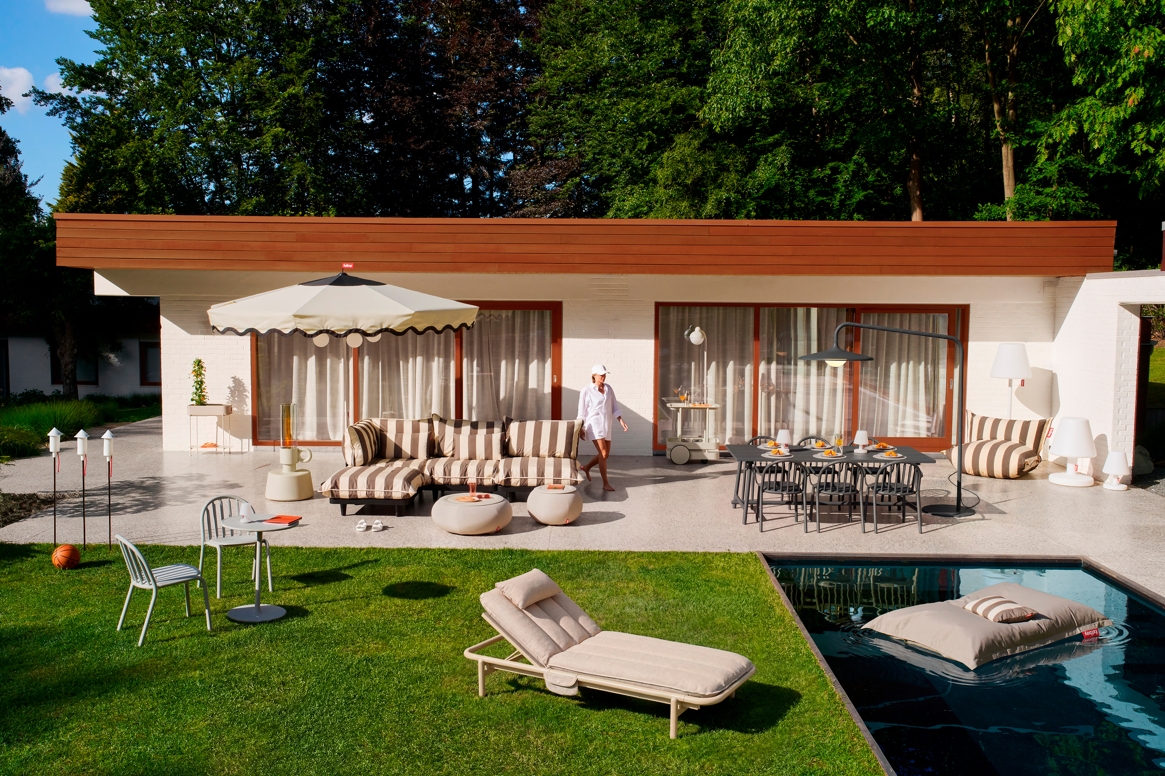 Patio moderne avec mobilier Fatboy incluant sofa Paletti, ensemble Fred's, parasol, chaise longue et pouf flottant dans la piscine.