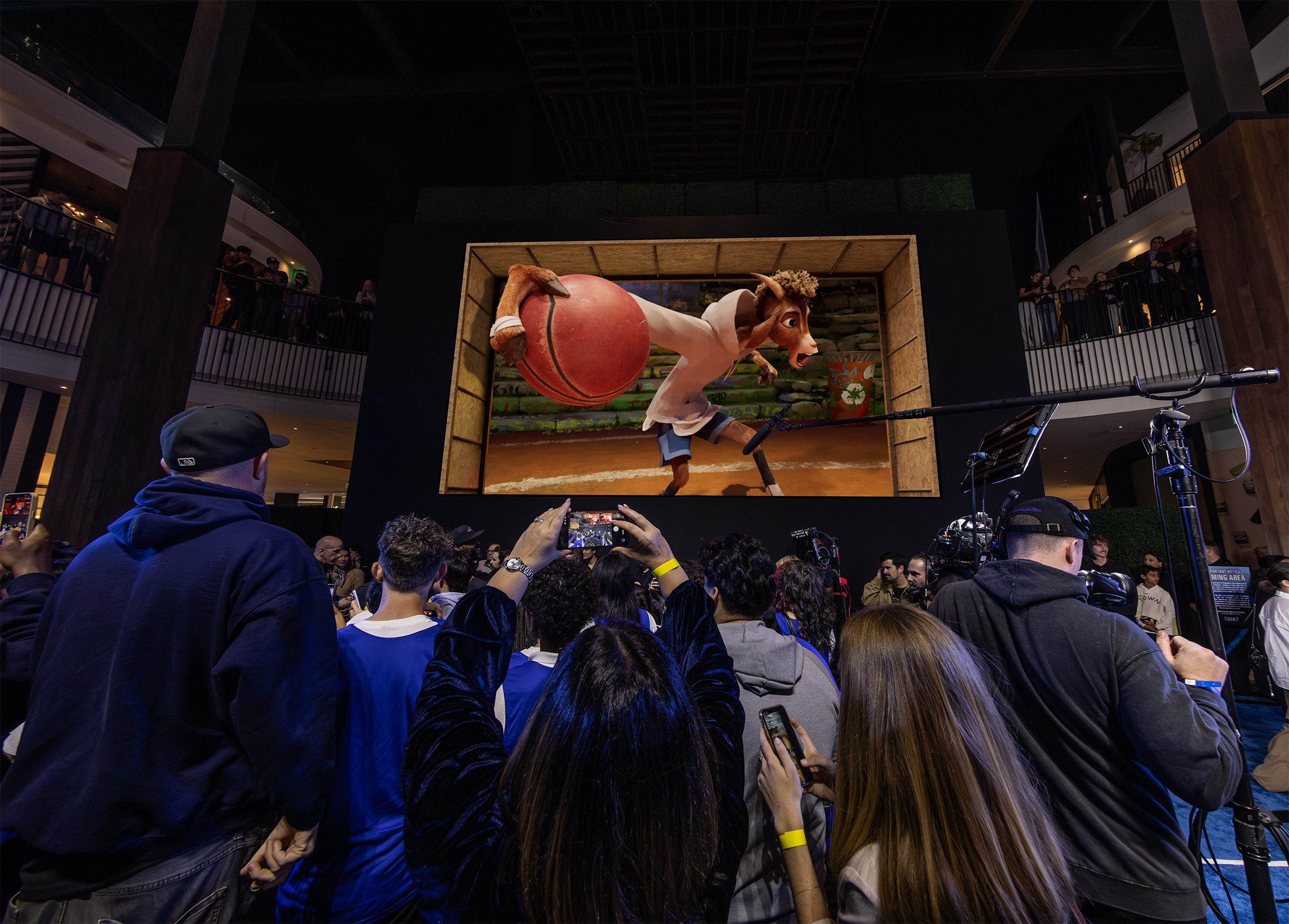Audience gathered in an indoor atrium watching a large-format 3D animated basketball scene on a digital display.
