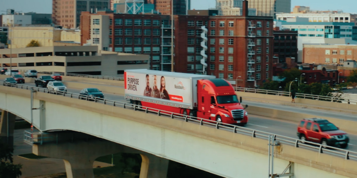 Red Nussbaum Transportation truck and Purpose Driven branded trailer on an elevated highway with city skyline in background.