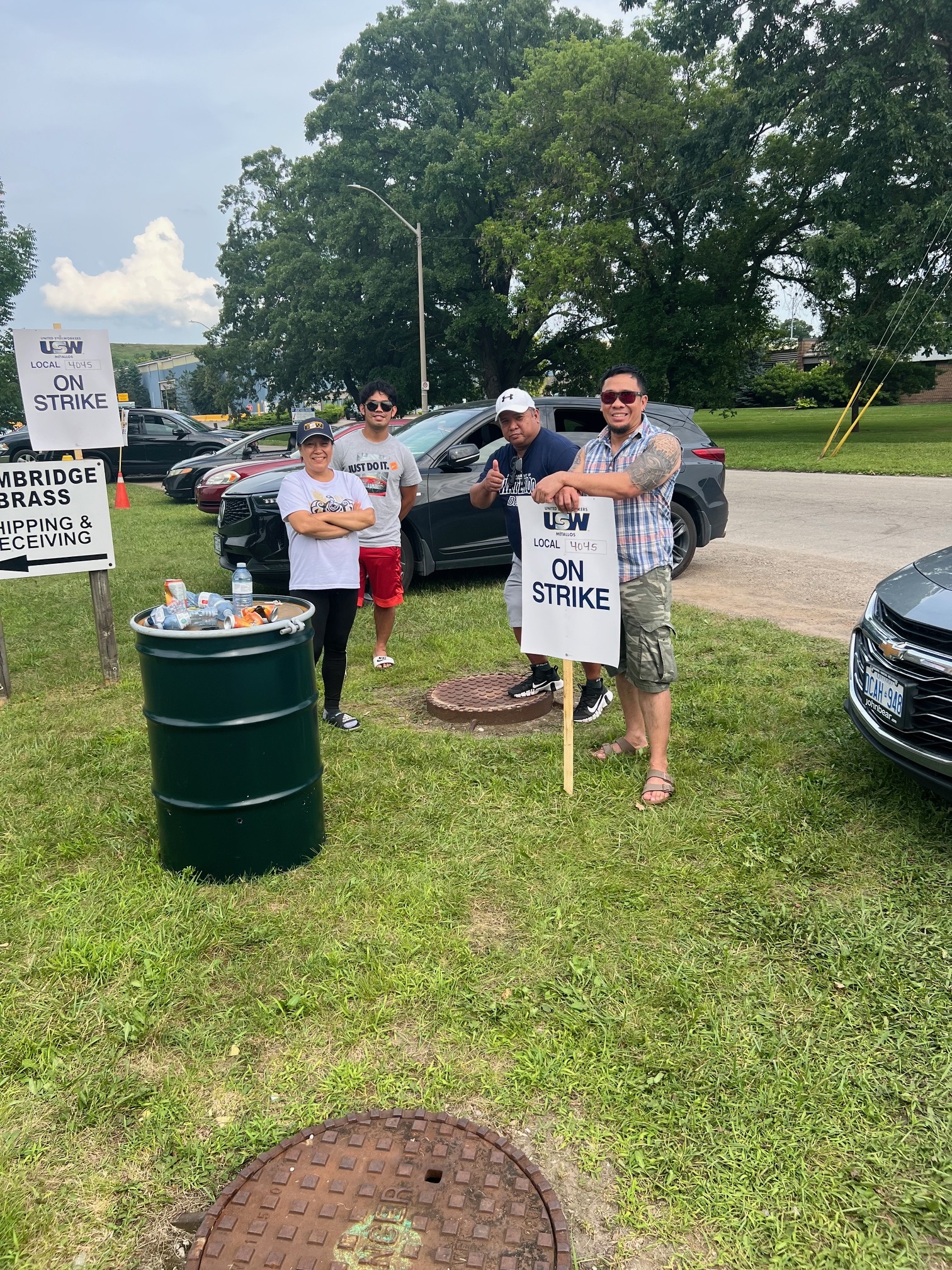 Image: Four people stand together outdoors on the grass, beside an oil drum, holding picket signs that say "On Strike."