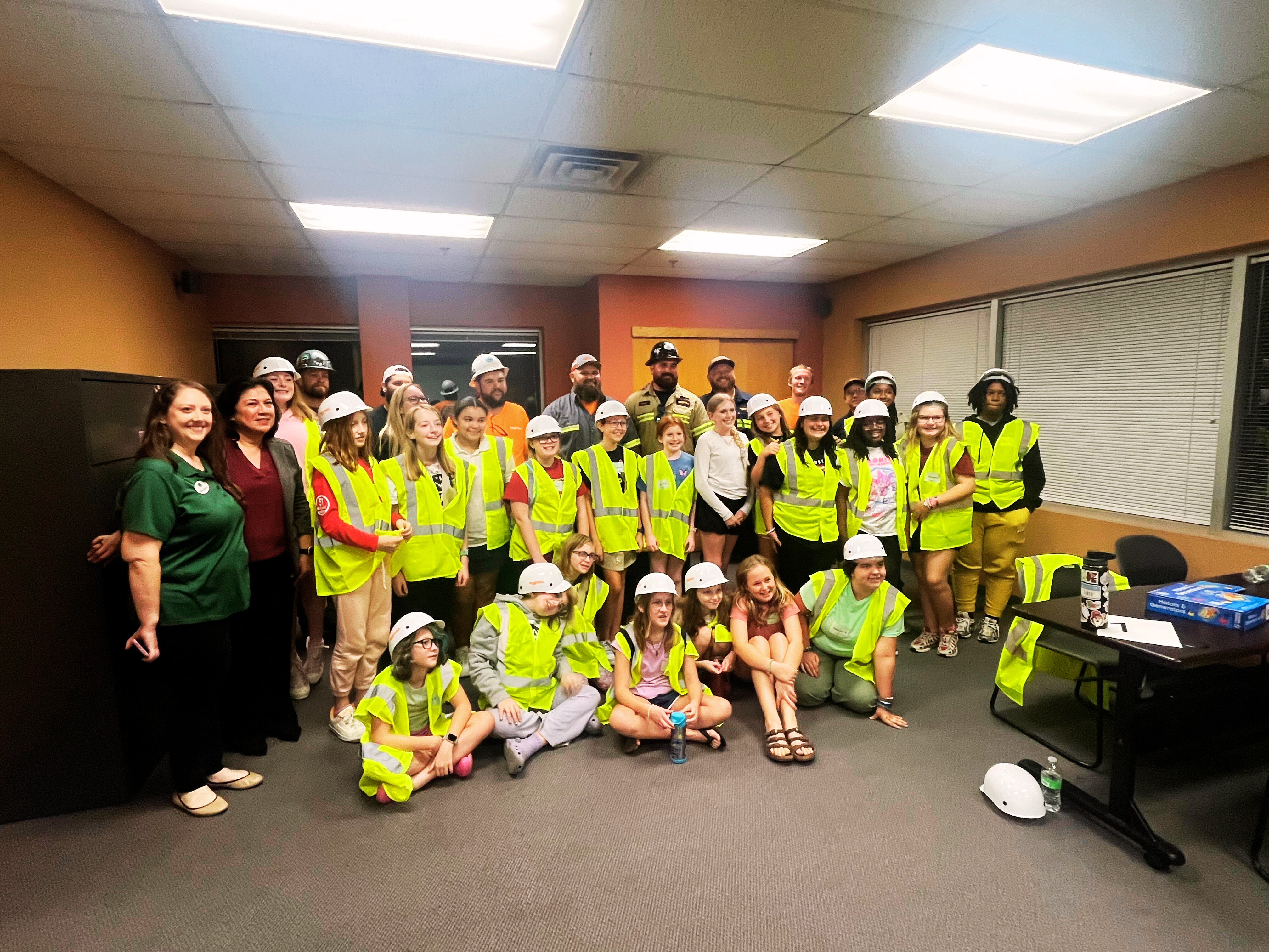A large group of children and adults pose together indoors. Most of them are wearing bright yellow safety vests and white hard hats