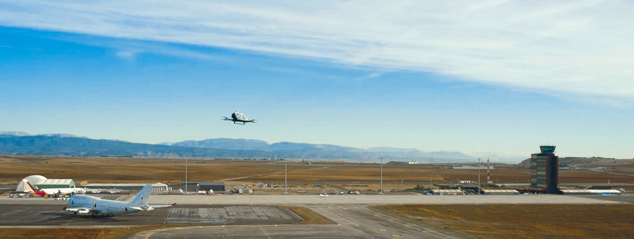 (Photo: EH216-S flying at LEDA with the airport, vertiport and planes behind)