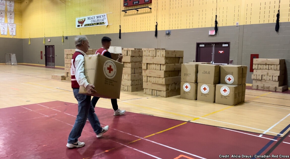 In a large gymnasium, two people carry large cardboard boxes away from the camera towards a stack of other large cardboard boxes.