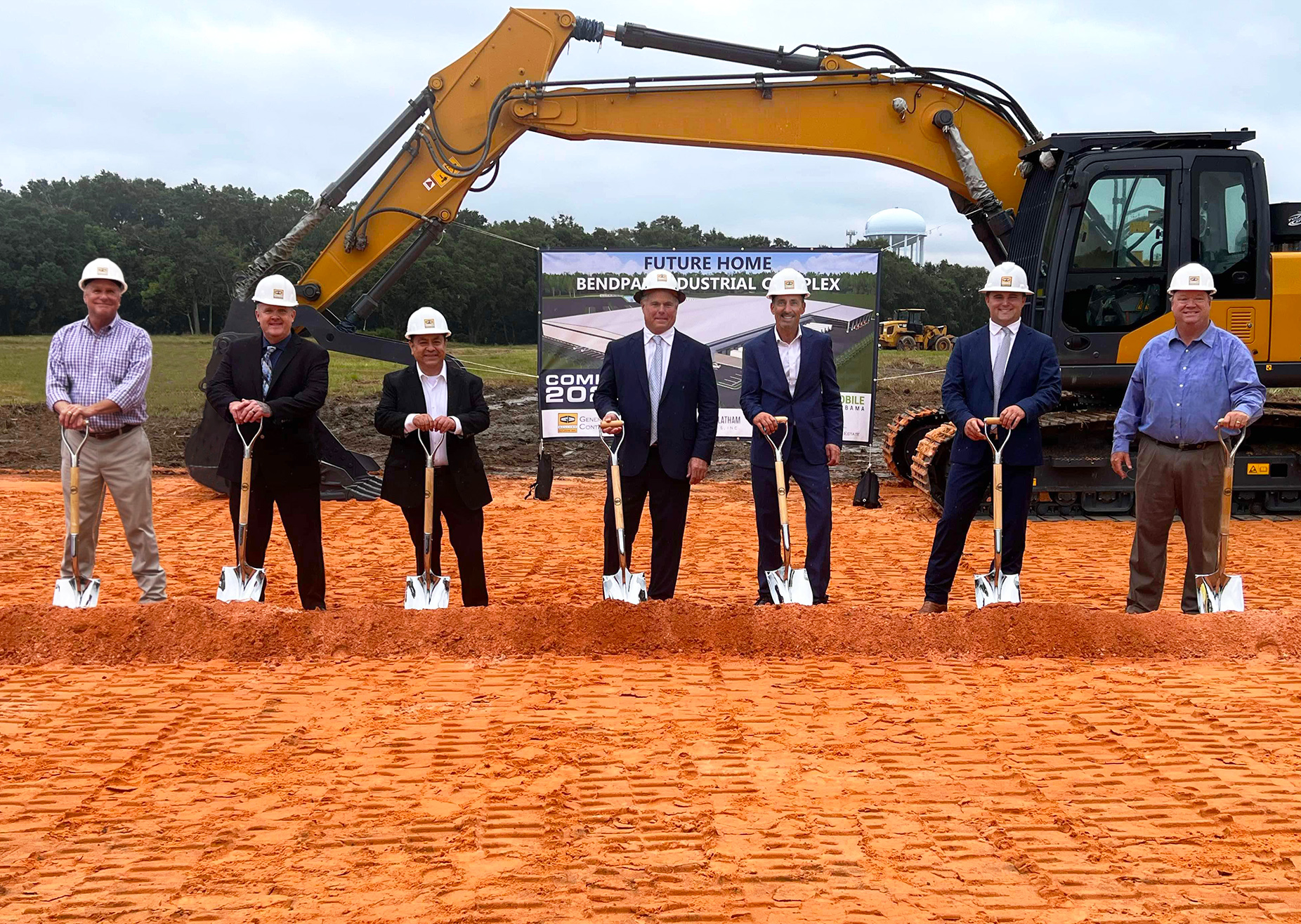 Men wearing hardhats and holding shovels stand in front of a large digger