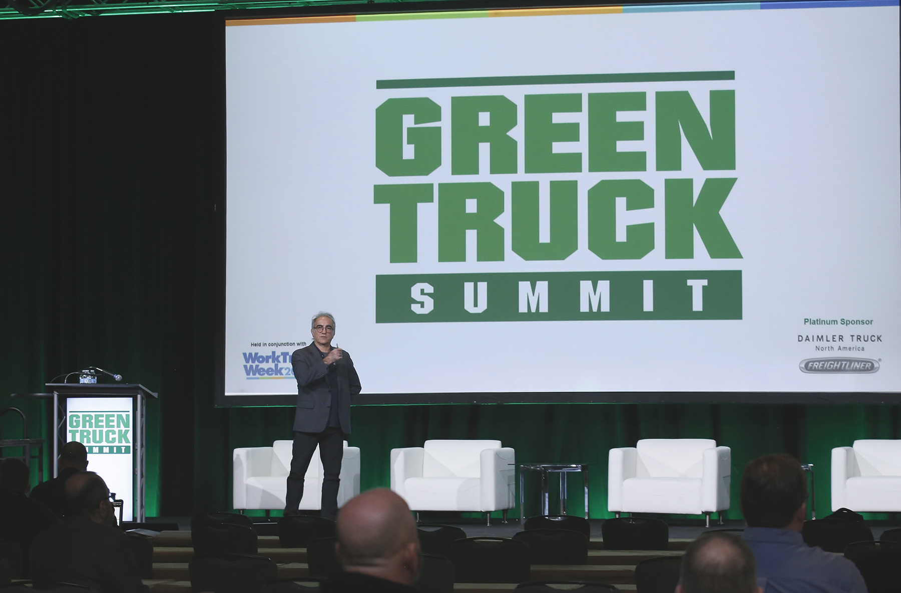 A male presenter stands on stage in front of a large screen that reads Green Truck Summit.