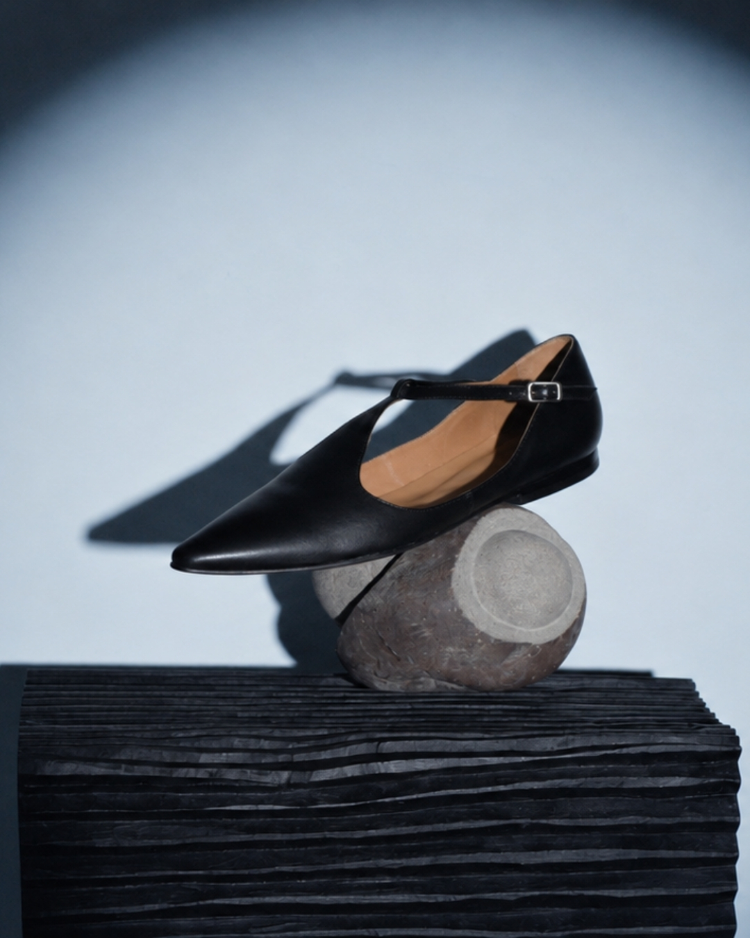 Black pointed T-strap flat displayed on a rounded stone form above a black textured pedestal.