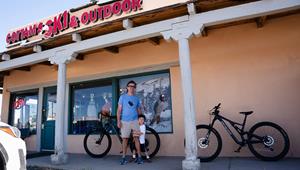 Images shows the exterior of the Cottam's Store in Downtown Taos with a father and son standing outside the store getting ready to go on a bike ride