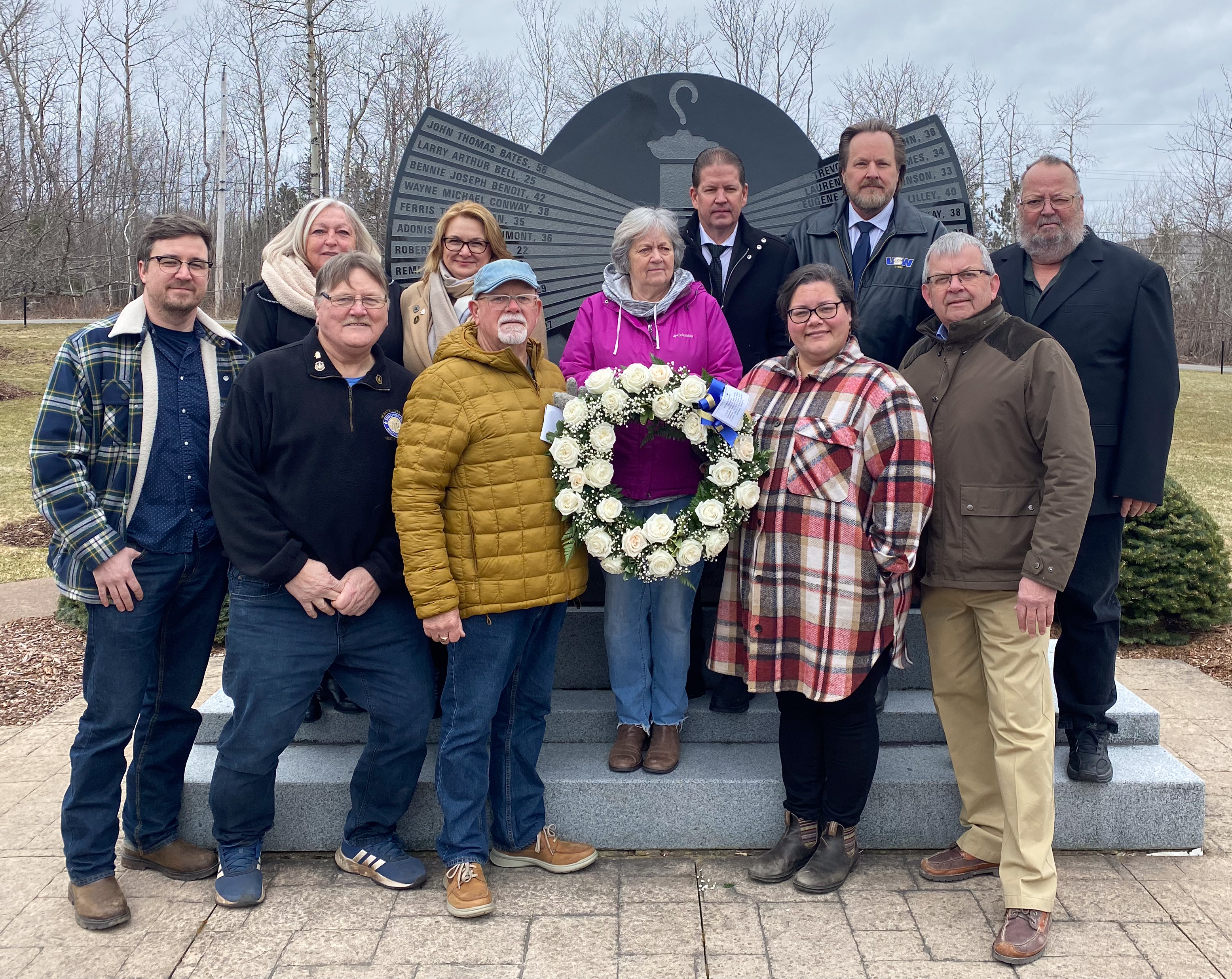 A group of 11 people is standing outdoors in front of a large granite monument. In the centre, one of the group holds a wreath with white roses.