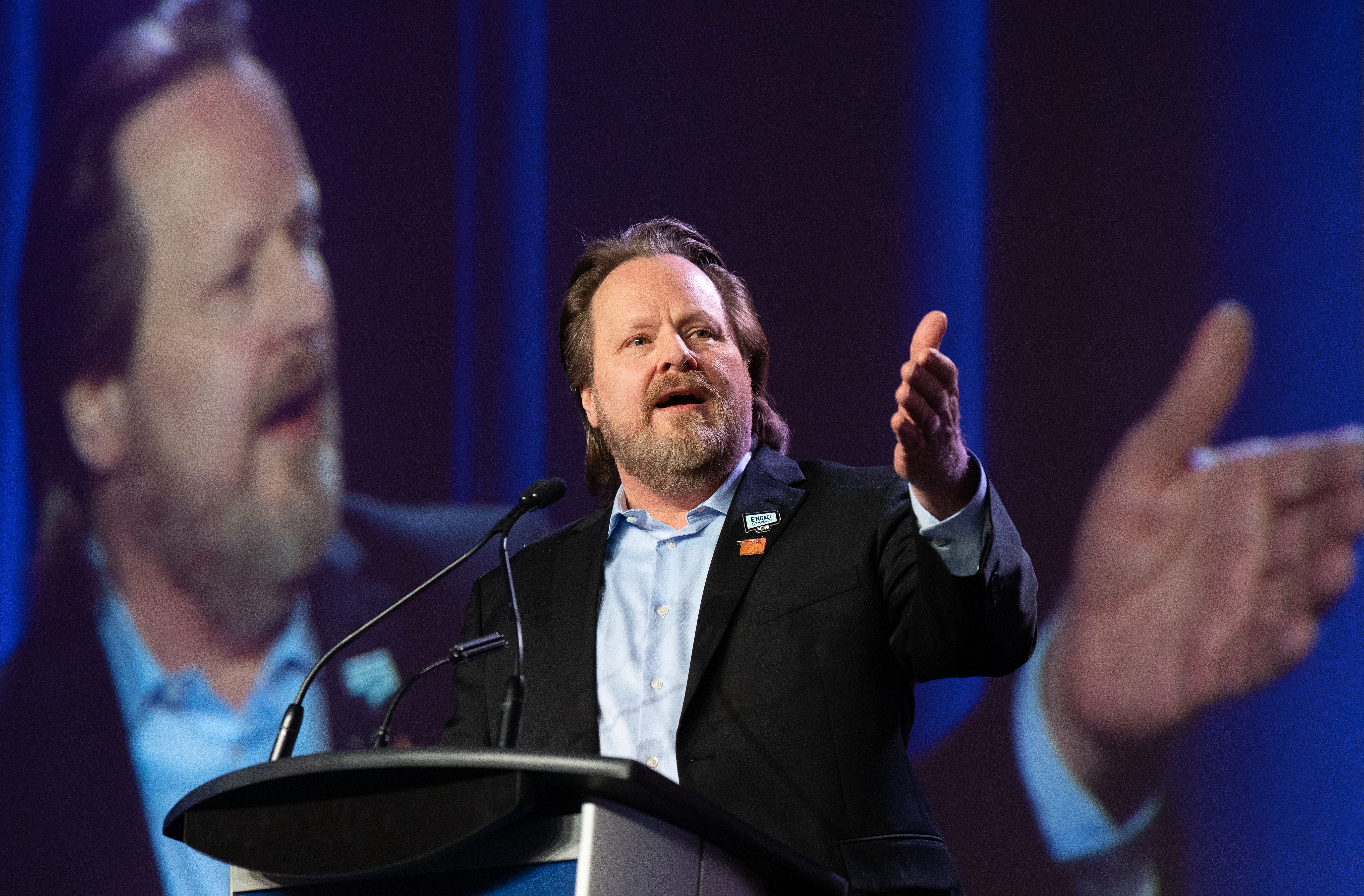 A bearded man in a jacket and shirt is at a podium speaking to an unseen audience at a conference event.