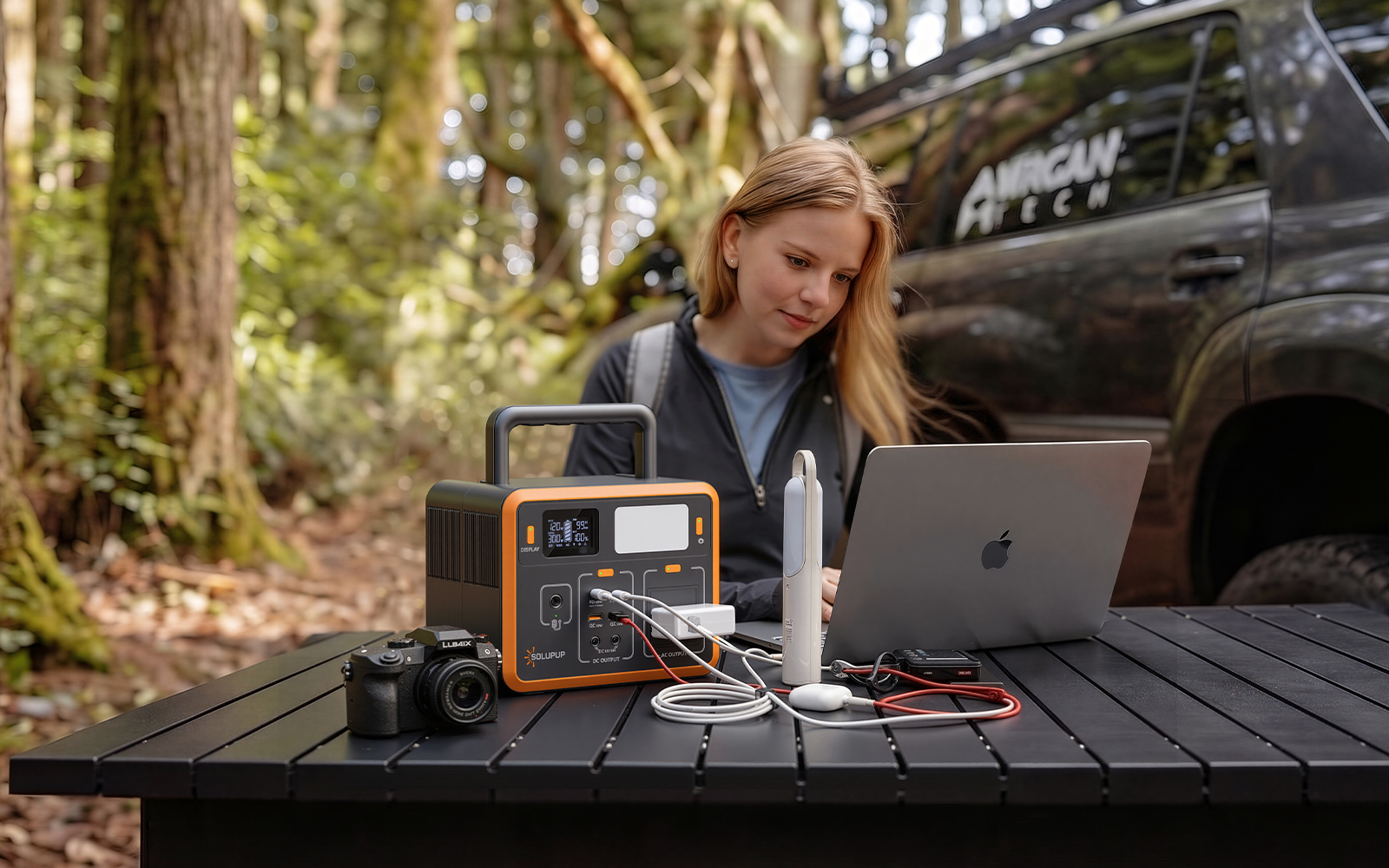 Portable power station charging laptop, camera, and devices outdoors on a table, ideal for camping or remote work in nature.