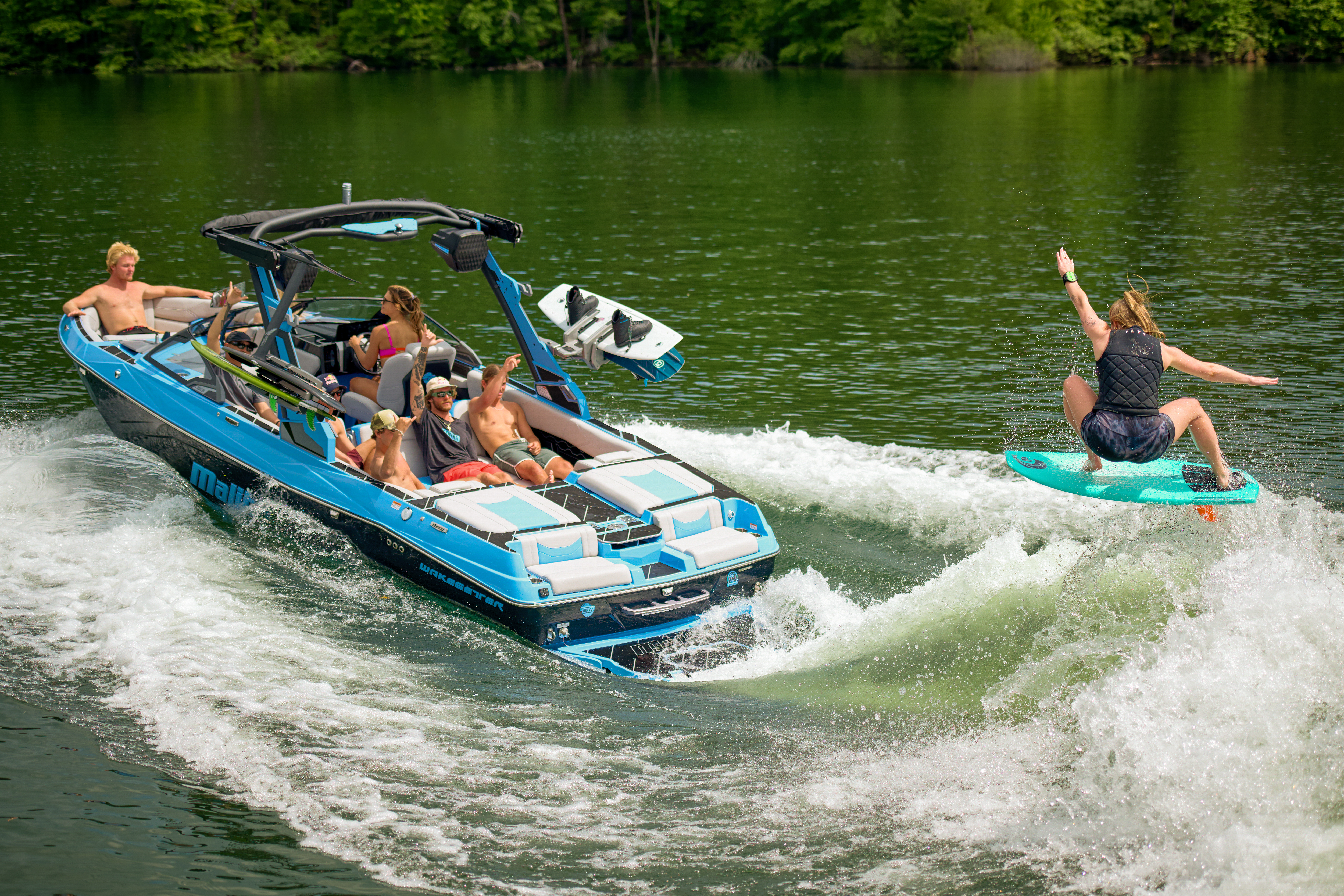 Wakesurfer Jordan Wolfe performs an aerial trick behind a blue Malibu Wakesetter 23 LSV on a calm lake, with friends onboard watching and cheering.