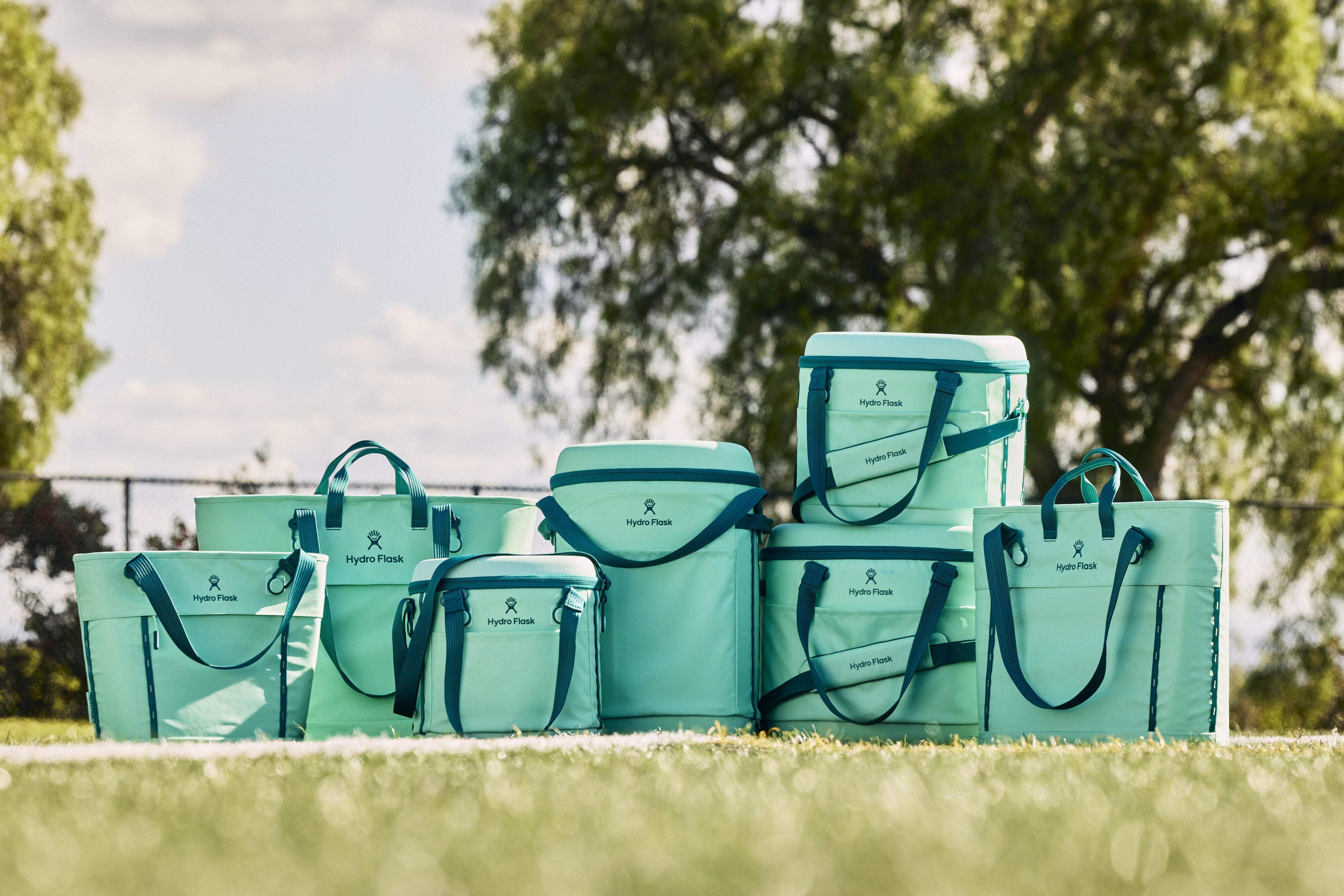 Seven Hydro Flask soft coolers in a vibrant mermaid green color are placed on a green field. A blue sky and green tree fill the background.