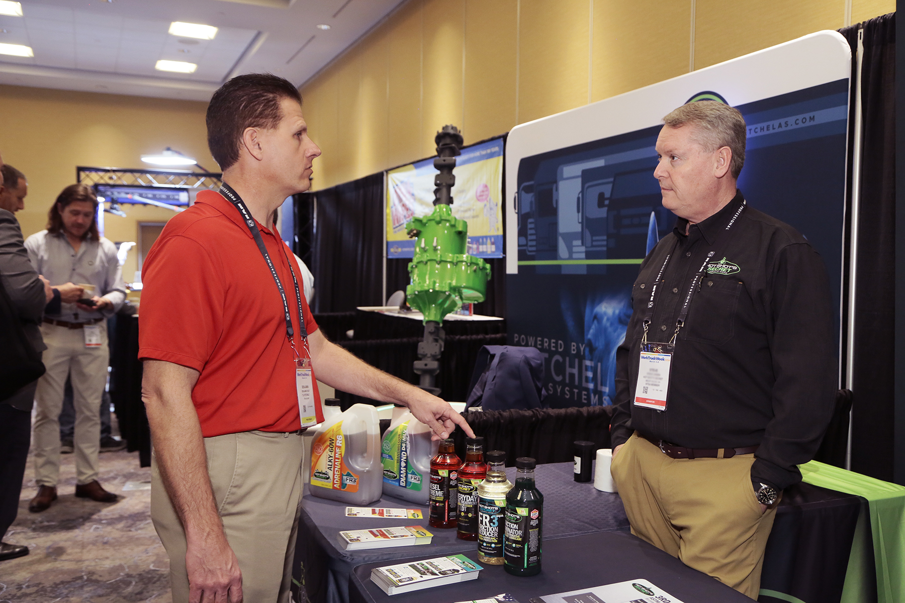 Two men discuss vehicle chemicals displayed on a table