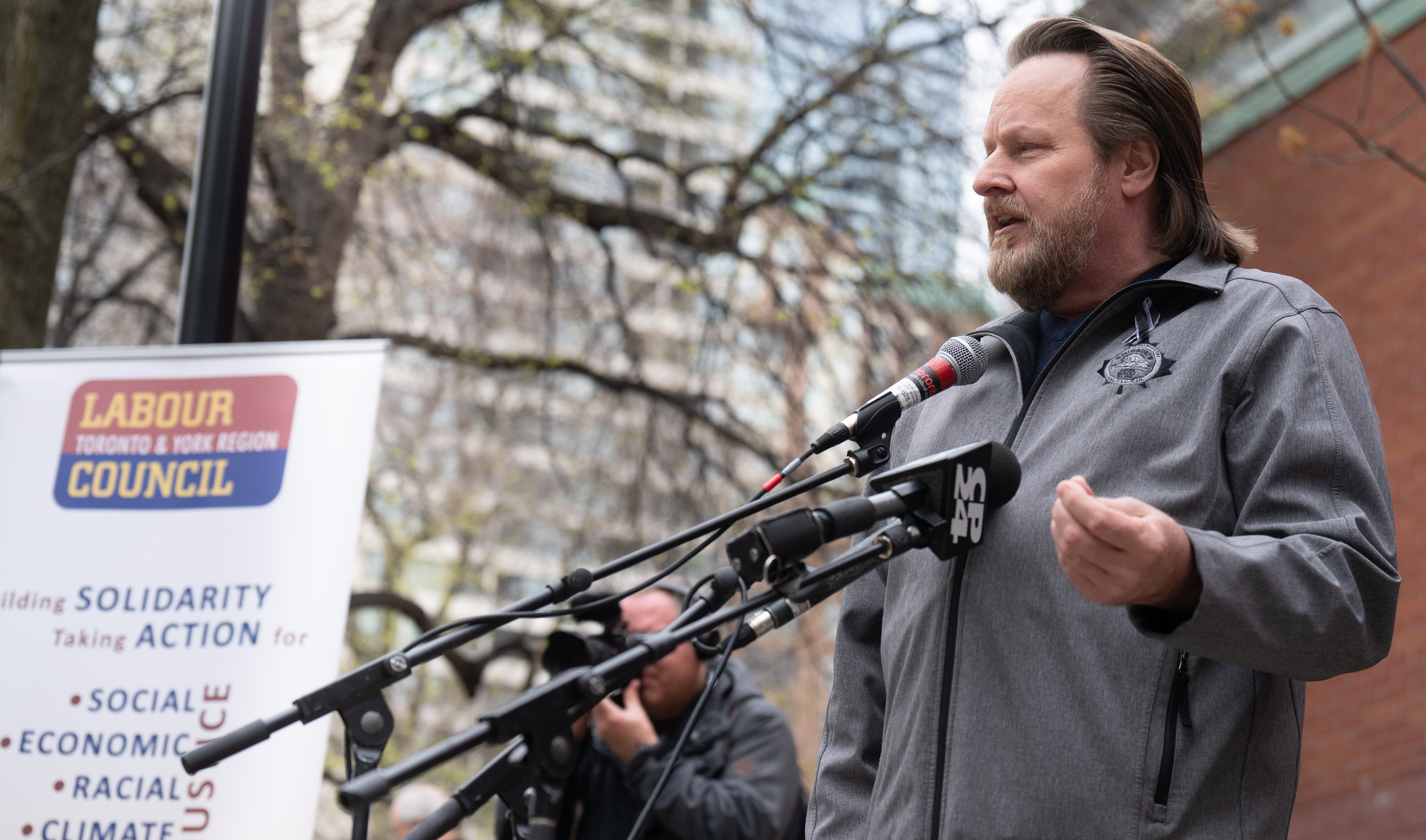 A bearded man in a grey jacket speaks at a micropohone at an outdoor labour event.
