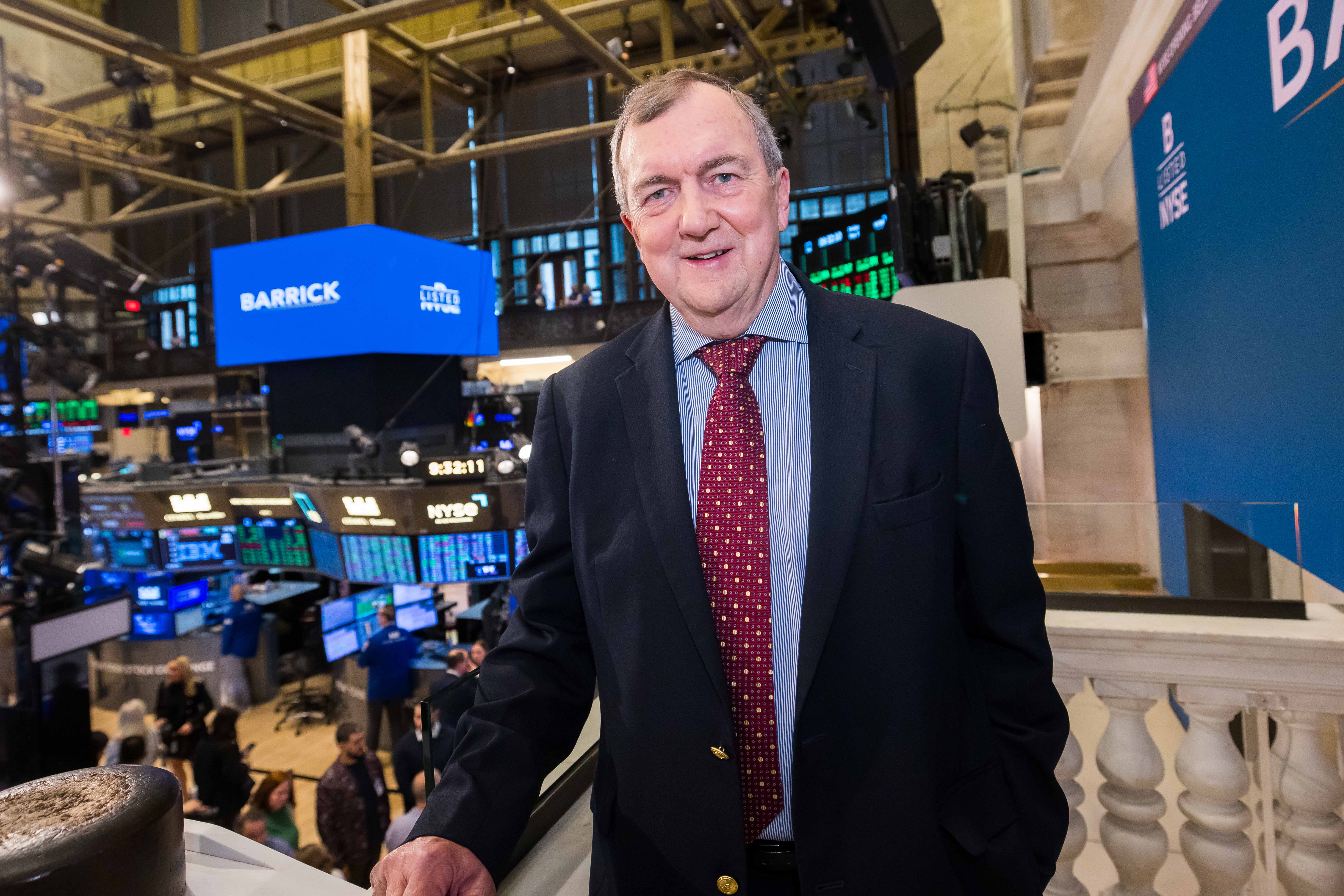 The image shows a man in a dark suit with a blue striped shirt and burgundy patterned tie overlooking the NYSE trading floor.