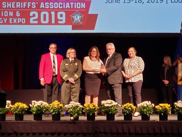 Ms. Delia Ginorio receiving the 2019 Crime Victim Services Award on behalf of the San Francisco Sheriff's Department at the NSA's annual conference. Pictured (L to R): Sheriff Vernon P. Stanforth, Secretary of the NSA Executive Committee; Captain Michele Fisher of the SFSD; Delia Ginorio, SFSD Rehabilitation Services Coordinator; NSA President Sheriff John Layton; Kathleen Krill of Appriss Safety.