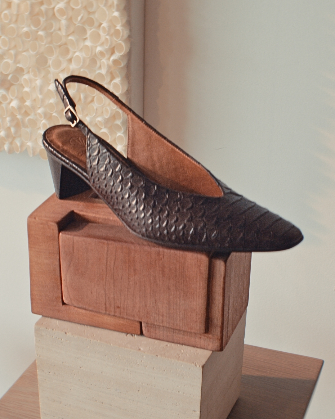 Dark brown snake-embossed slingback pump with a pointed toe and low sculpted heel displayed on stacked wooden forms.
